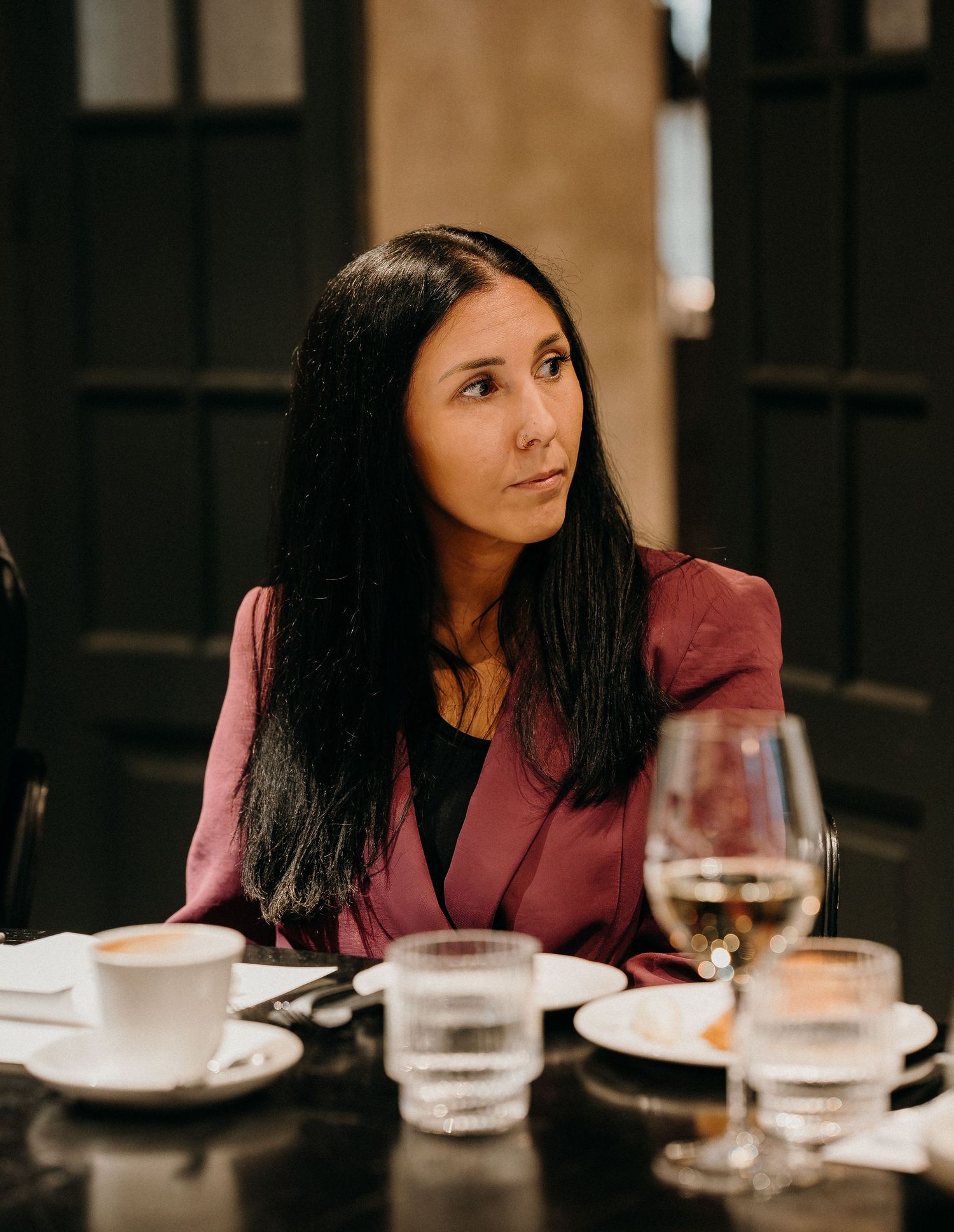 A woman in a burgundy top sits at a dining table with wine glasses and plates during a meal.