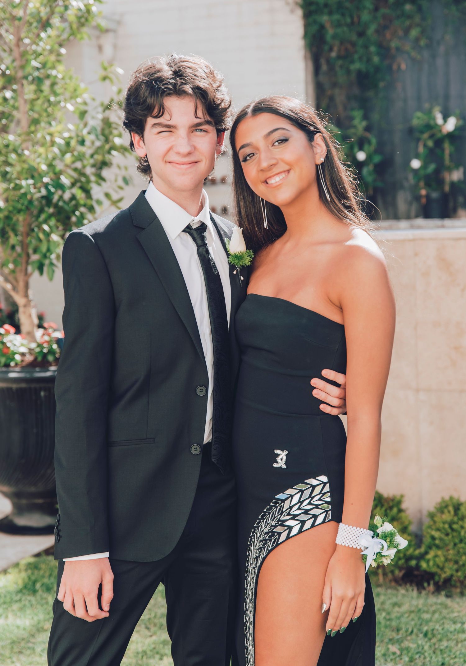 A formal couple in classic black attire stand close together in an outdoor garden setting against greenery.