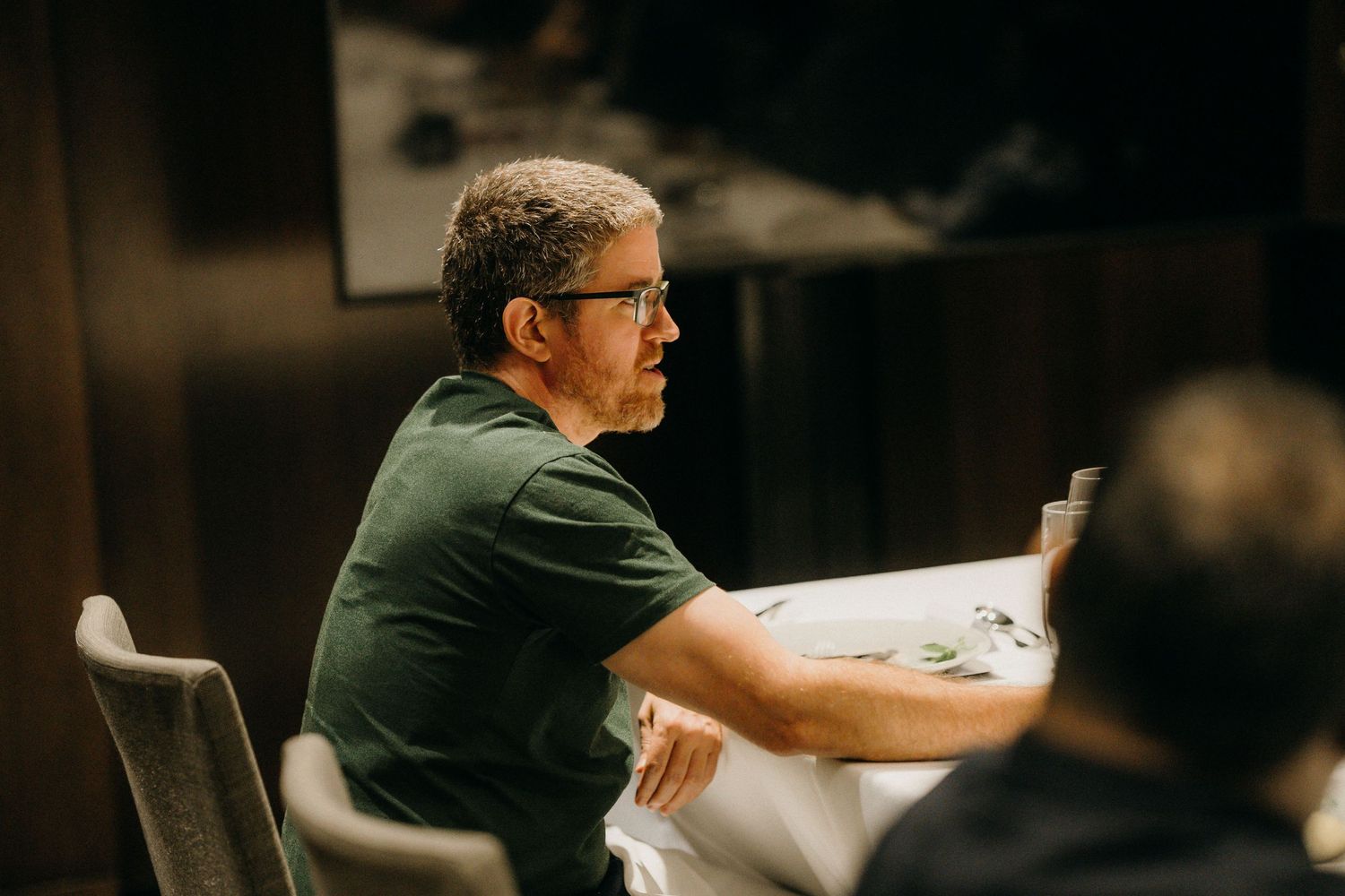Middle-aged man with glasses in green t-shirt sitting at a restaurant table, looking thoughtfully to the side.