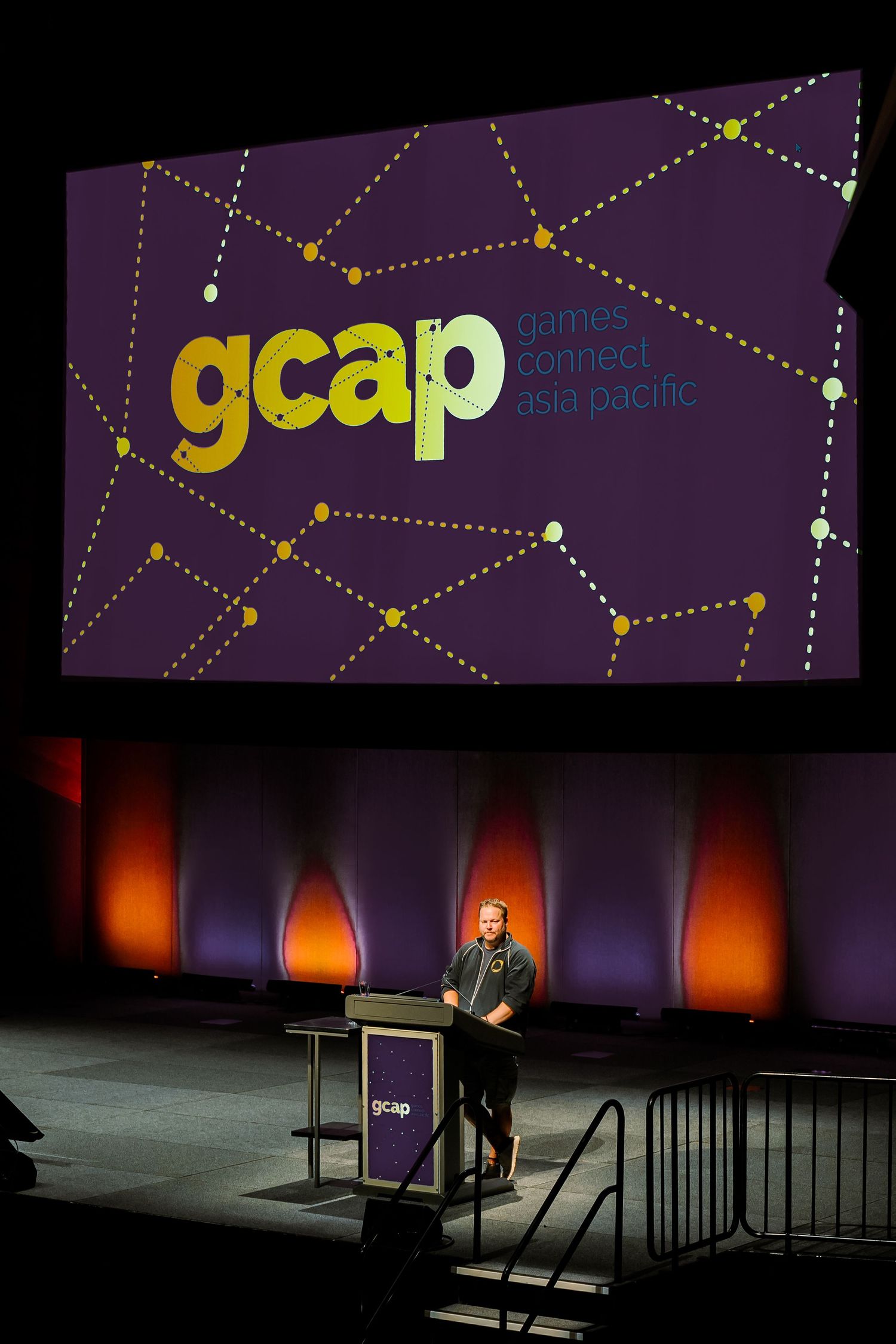 A stage presentation with purple lighting showing the GCAP logo on screen while a speaker stands at a podium below.
