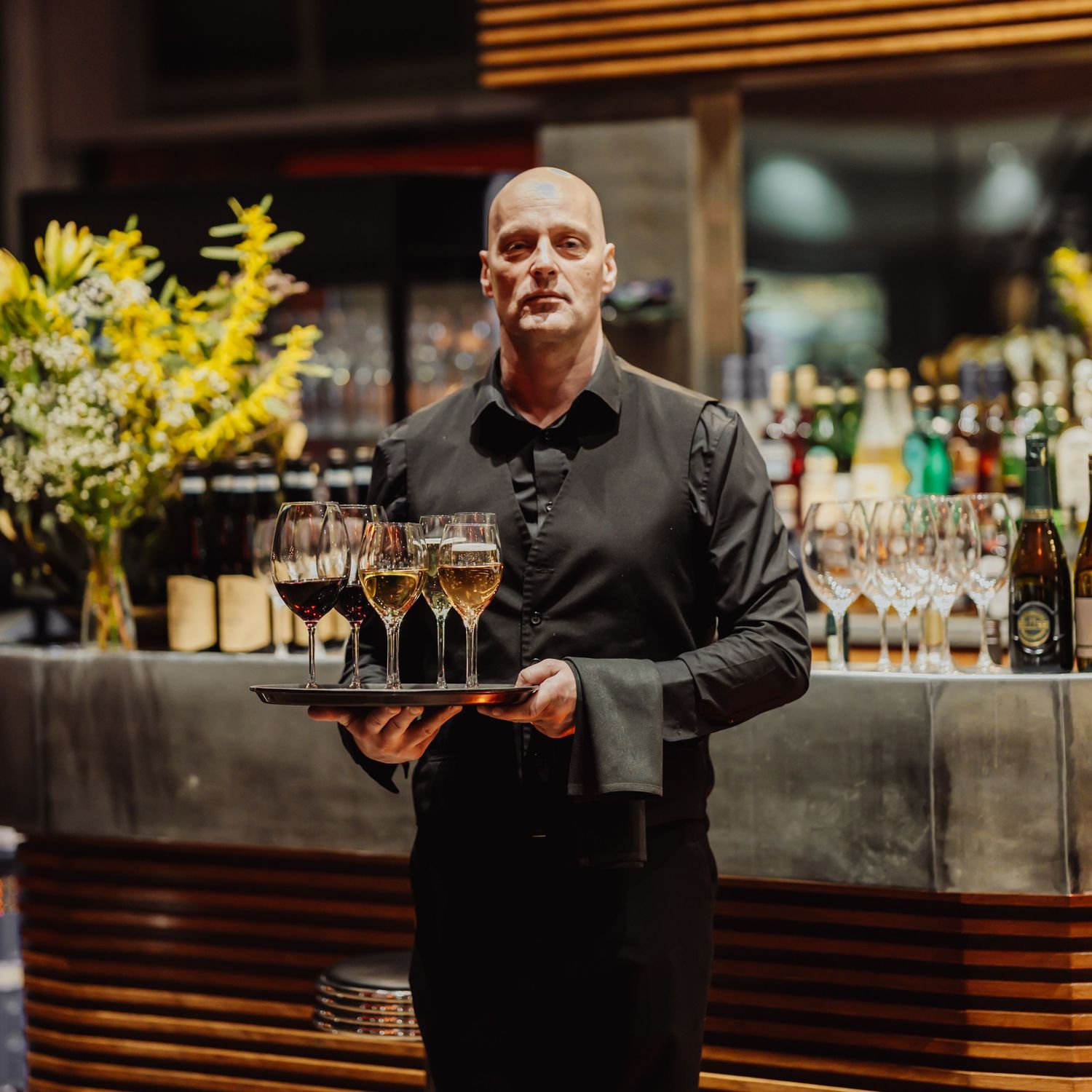 A restaurant server in black attire carries a tray of wine glasses past a bar with yellow floral arrangements.