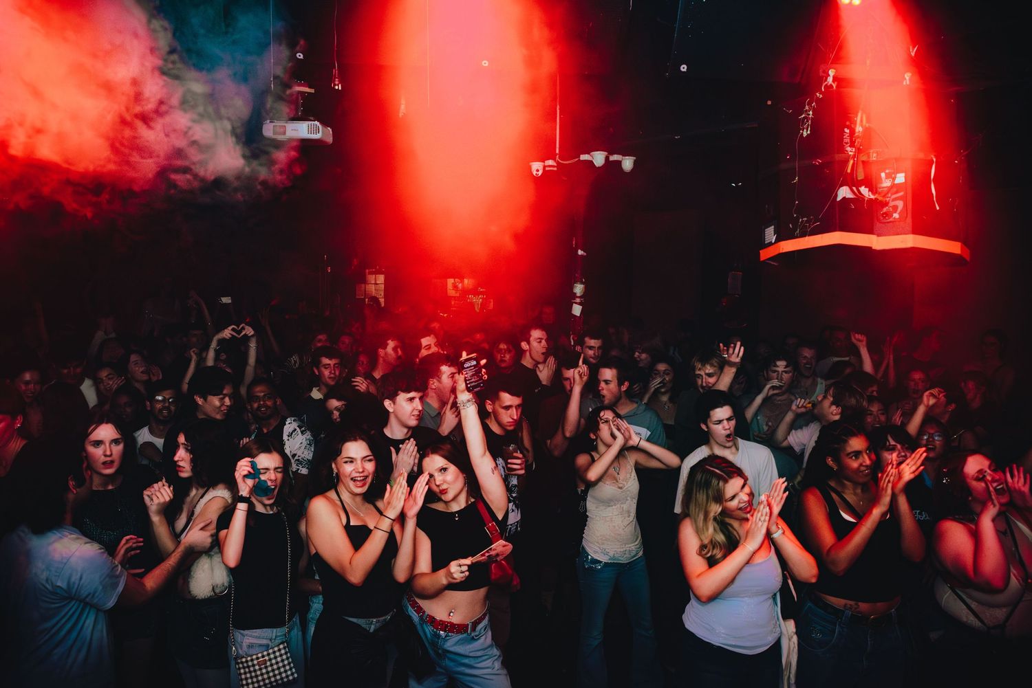 Crowd dancing at a nightclub with red lighting and atmospheric smoke effects filling the dark space.