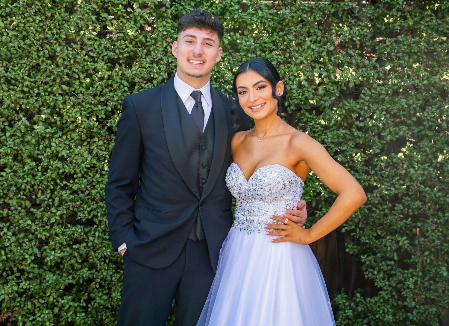A couple poses in formal attire in front of a lush green hedge wall at a special event.