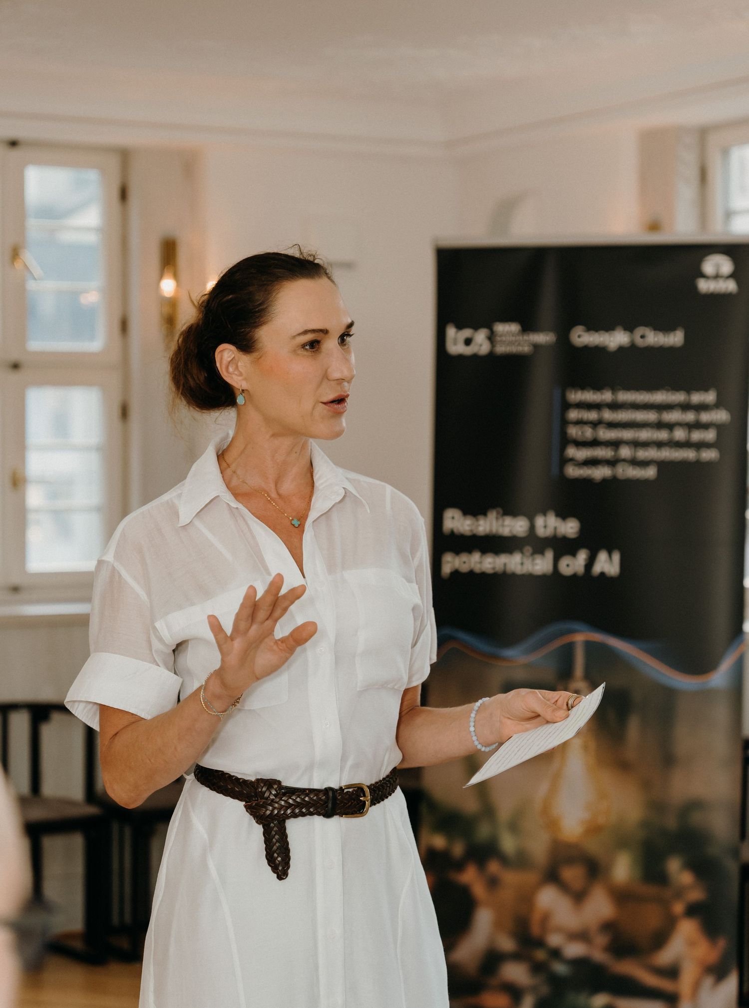Woman in white dress presenting at TCS and Google Cloud AI event, holding notes and gesturing confidently.