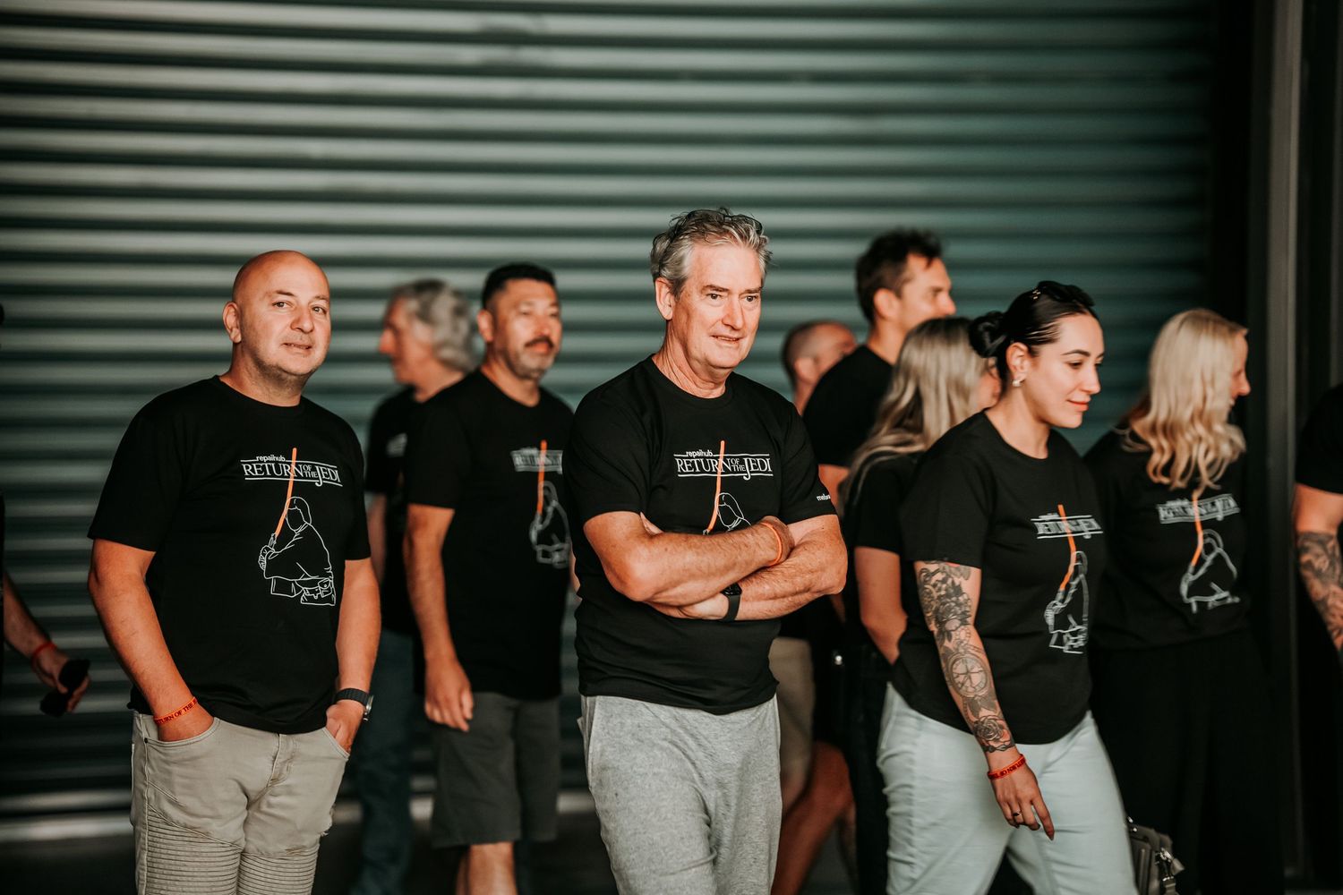 A group of people wearing black t-shirts pose together against a corrugated metal wall in a casual setting.