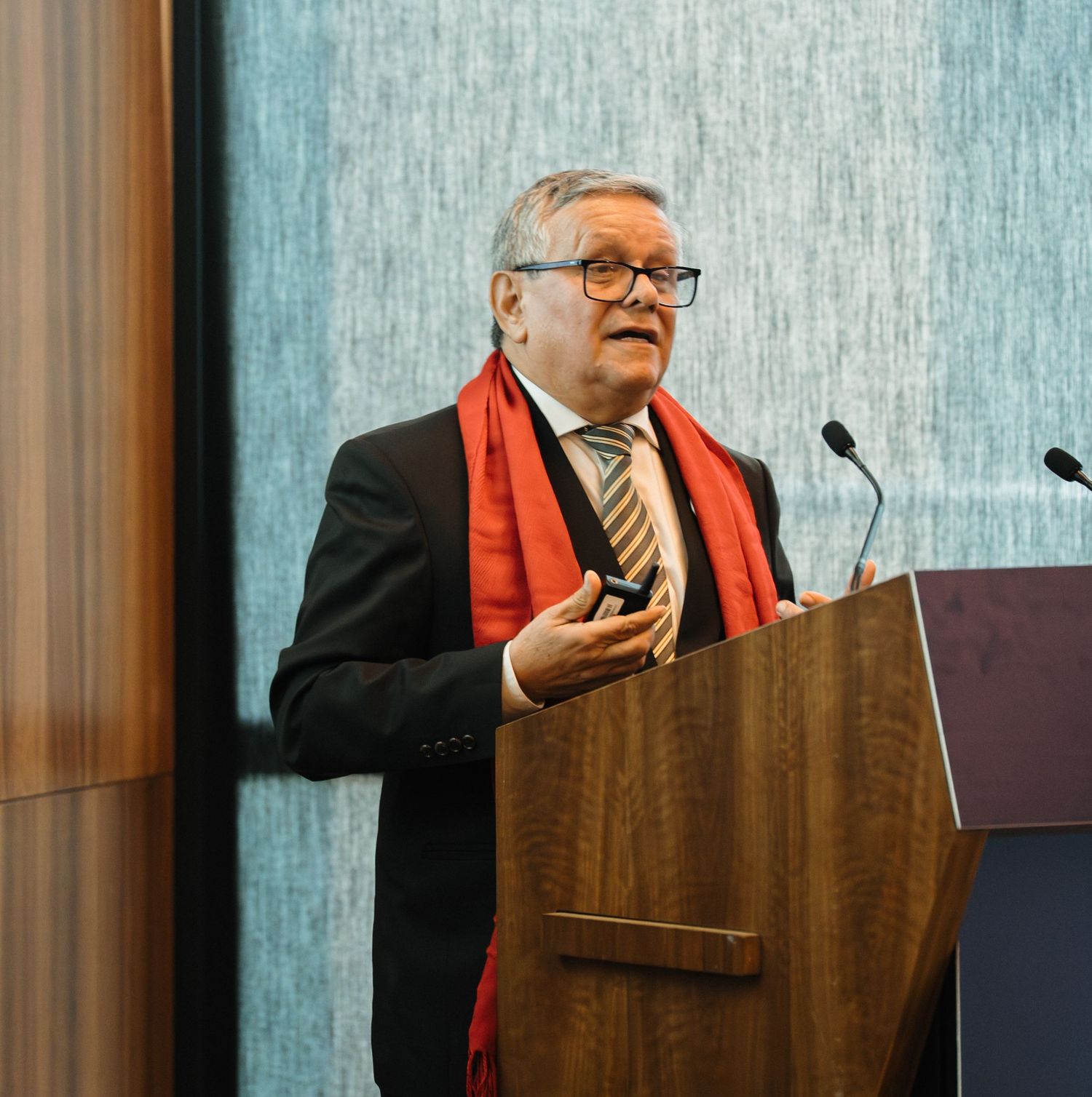Speaker at a podium wearing glasses and a red scarf gestures while giving a presentation at a conference.