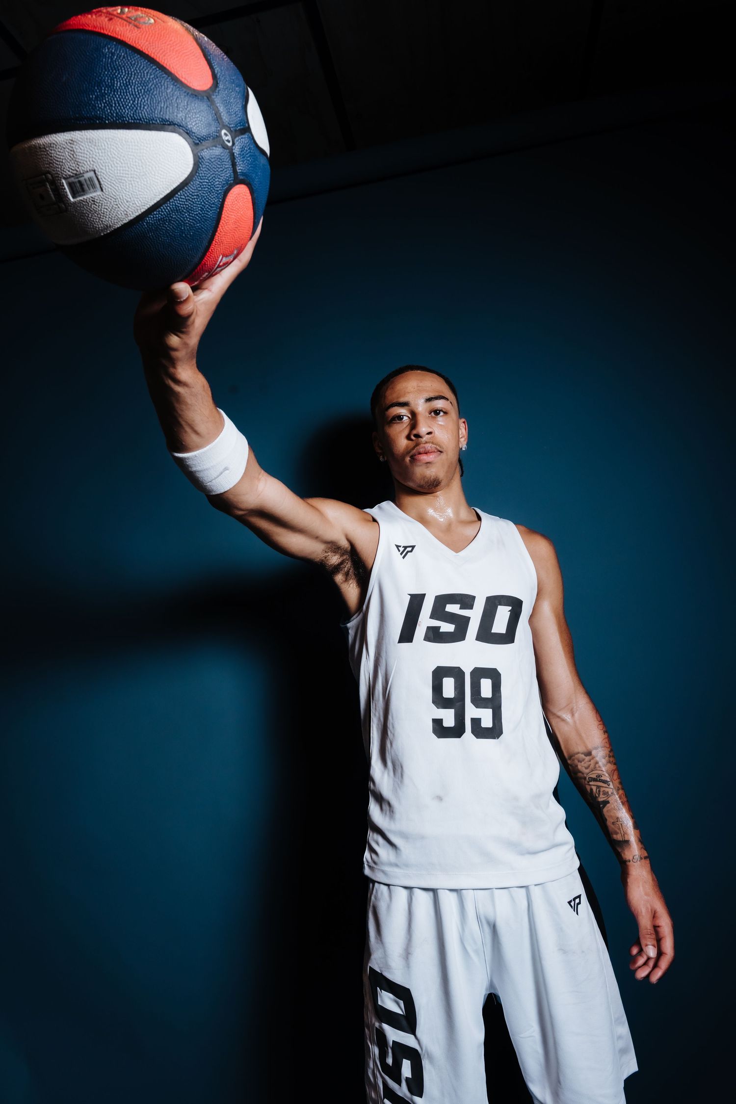 Basketball player in white ISO 99 uniform holds up a red and blue ball against dark background in dramatic lighting.