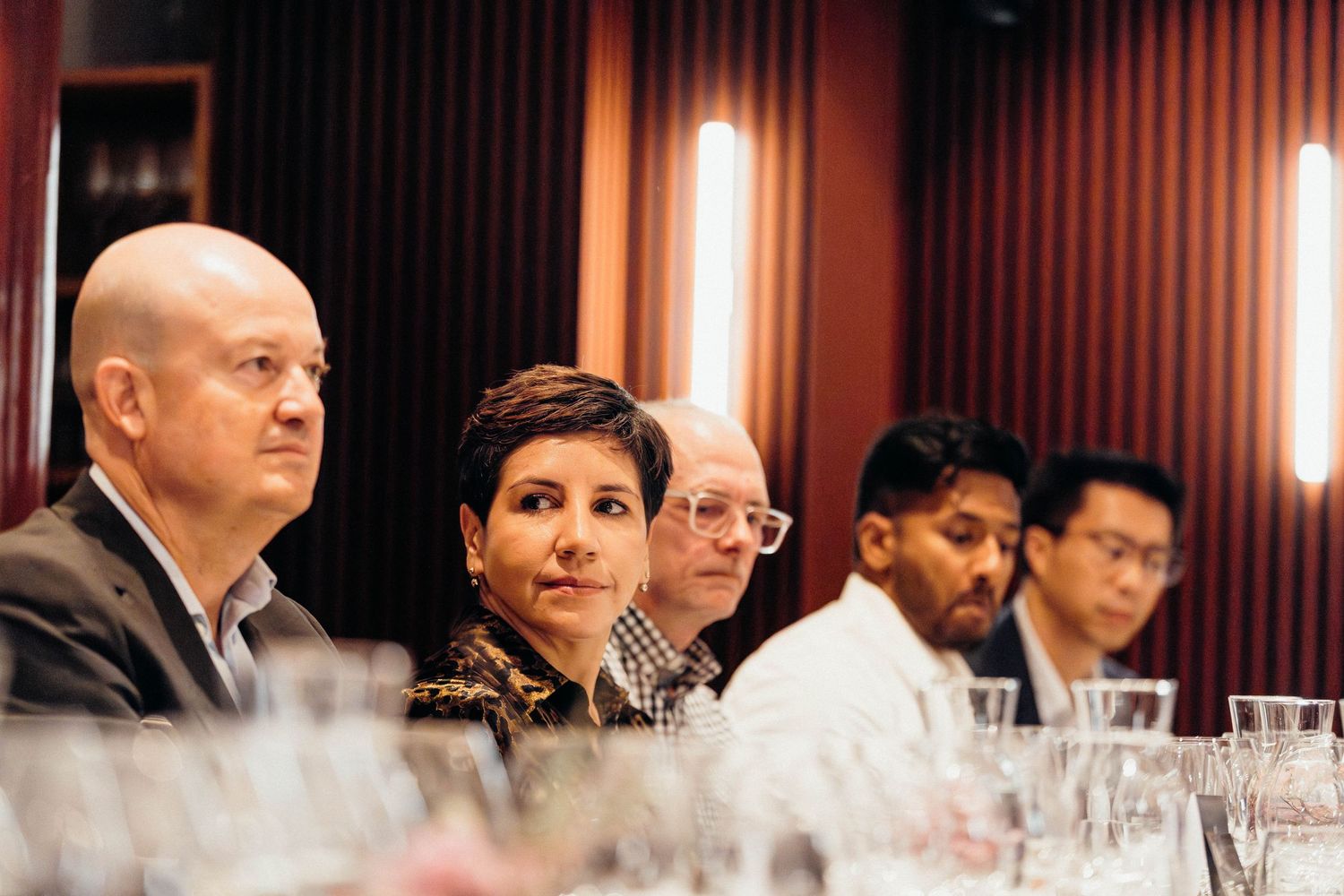 People seated at a formal event with wine glasses on the table and warm lighting in a wood-paneled room.