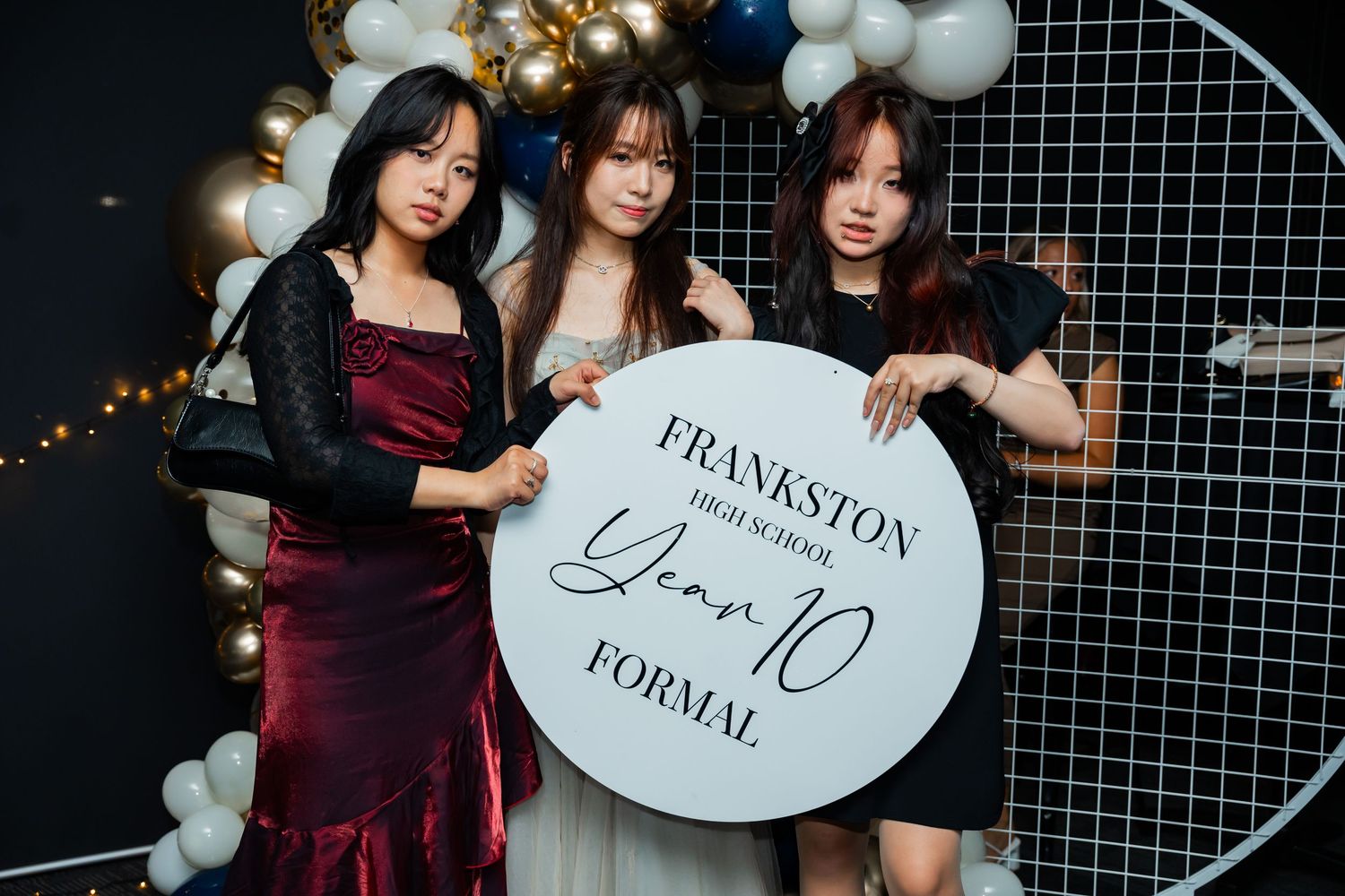 Three people in formal attire pose with a round sign at a formal event with balloon decorations in the background.