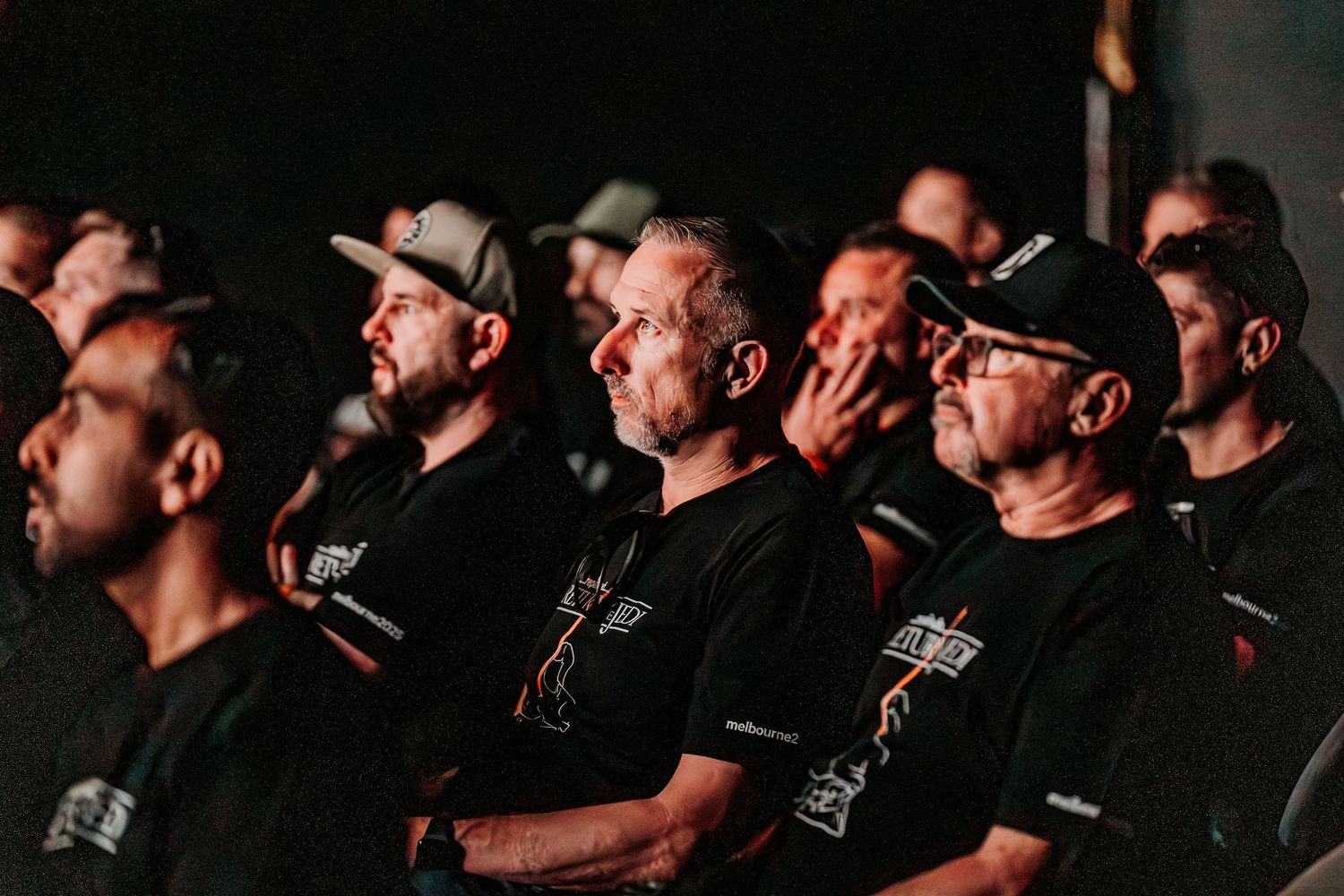 A group of people wearing matching black t-shirts sit together in a dark audience looking attentively at something off-camera.