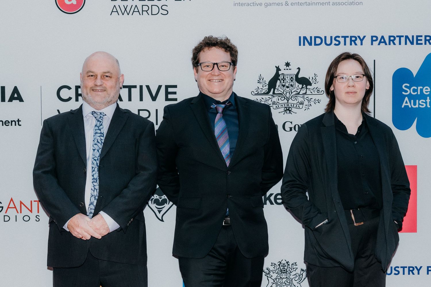 A group of people in formal attire standing in a row at an awards ceremony with sponsor logos visible behind them.