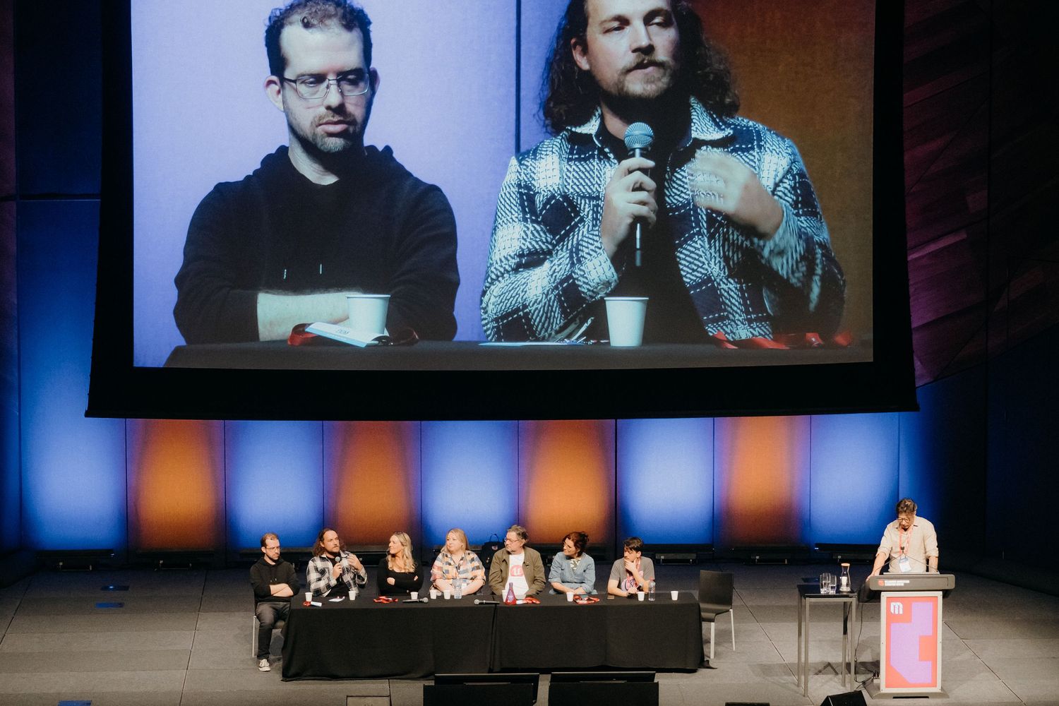 Panel discussion on stage with large projection screen showing two speakers above in theatrical setting.