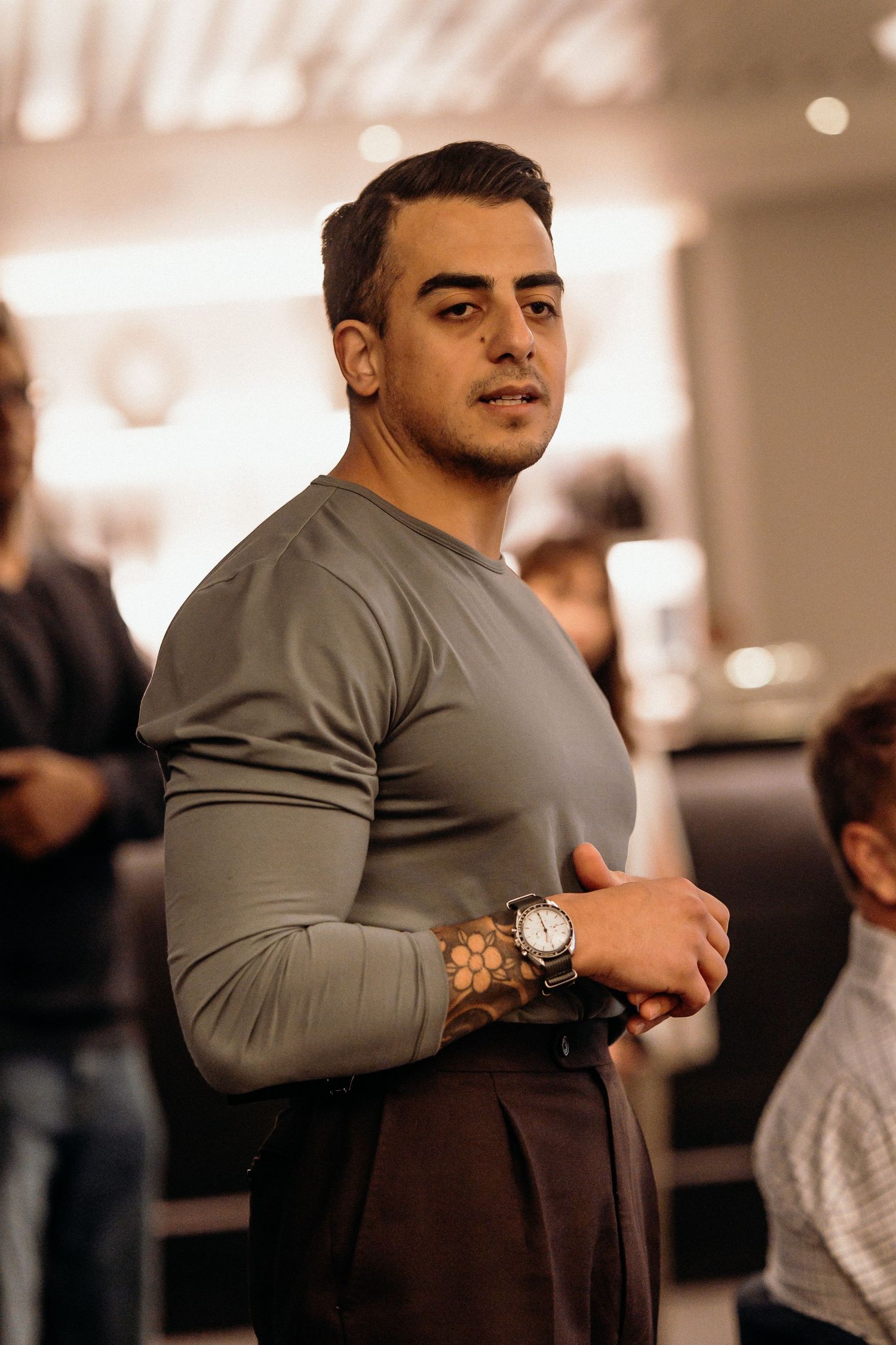 A person in a gray sweater adjusts their sleeve while standing in a dimly lit room with bokeh lights in background.