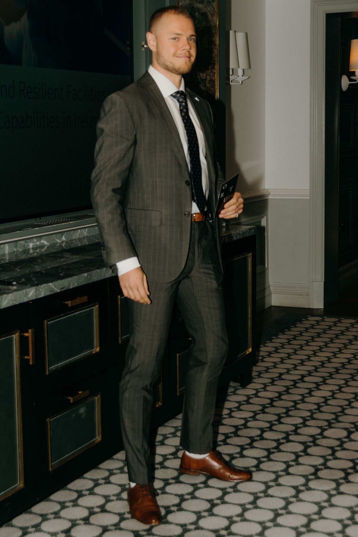 A person in a grey suit stands on a geometric patterned floor in a dimly lit hallway.