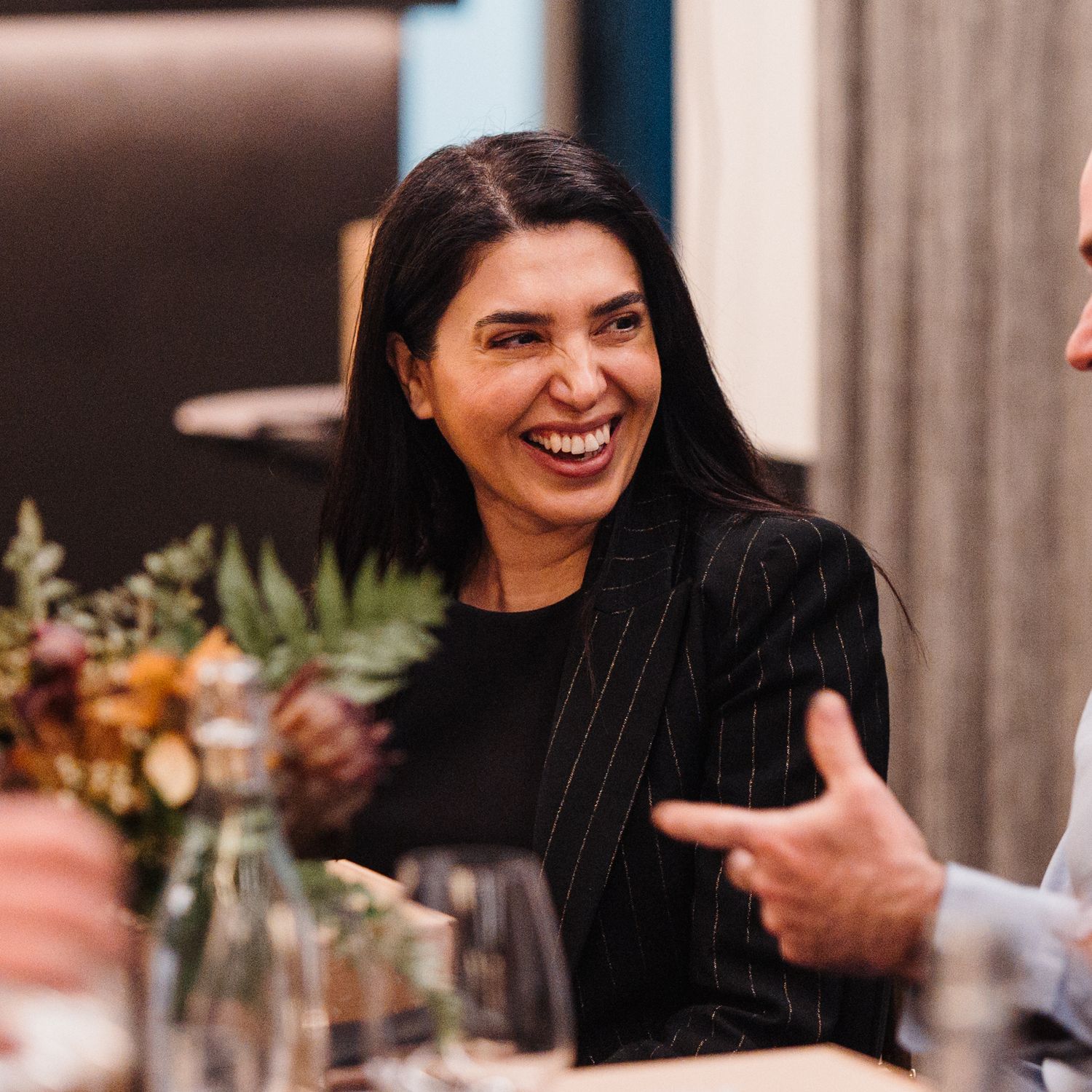 A person with dark hair smiling and laughing while sitting at a dining table with decorative place settings.