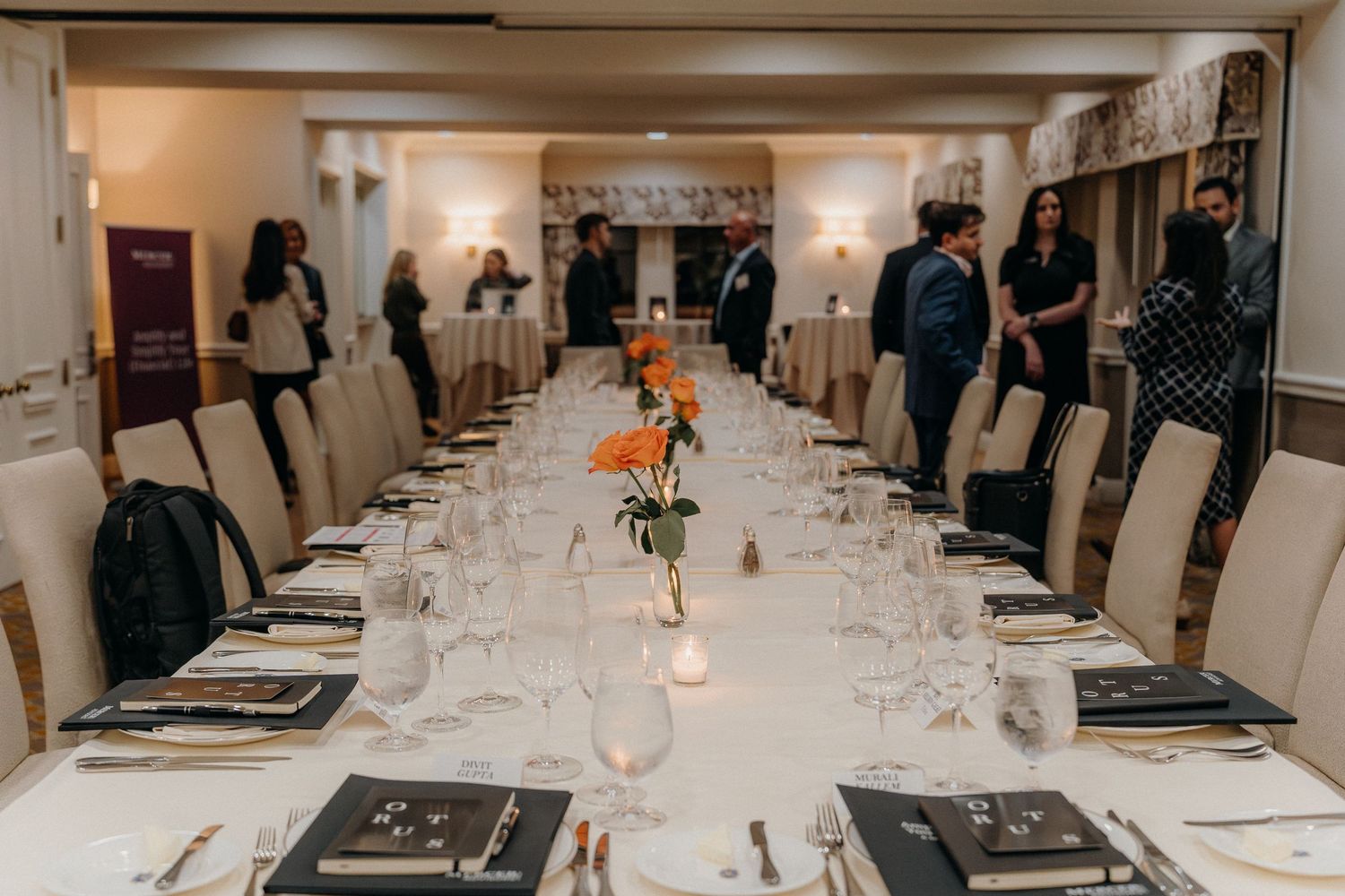 An elegant long dining table set for a formal dinner with white linens, place settings and orange floral centerpieces.