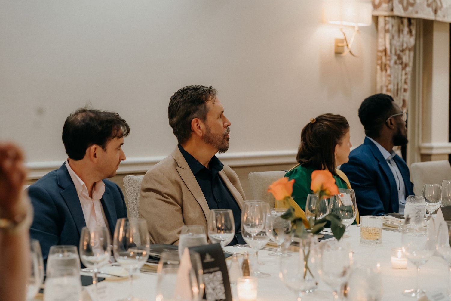 Guests seated at an elegant dinner table with wine glasses and candlelight in a formal dining room.