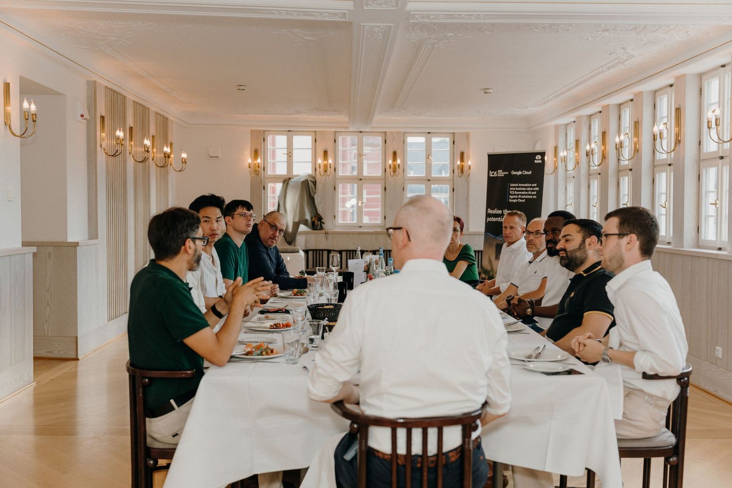 Group of people dining together at a long white-clothed table in an elegant bright restaurant with wall sconces.
