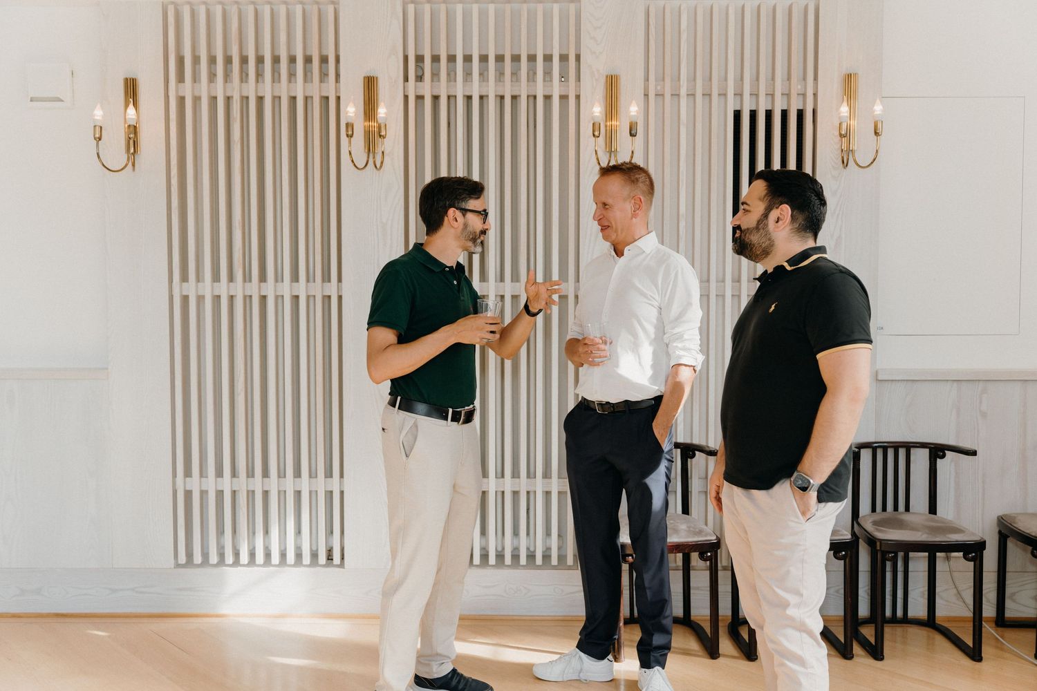 Three men in smart casual attire engaged in conversation in a bright, elegantly decorated white interior space.