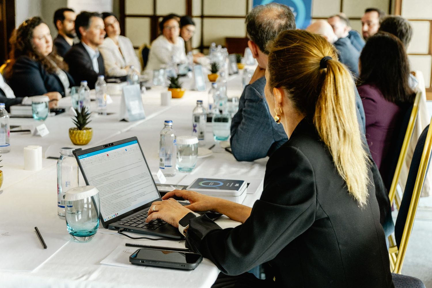 Professional woman using laptop at a corporate conference meeting with business attendees seated at a long table.