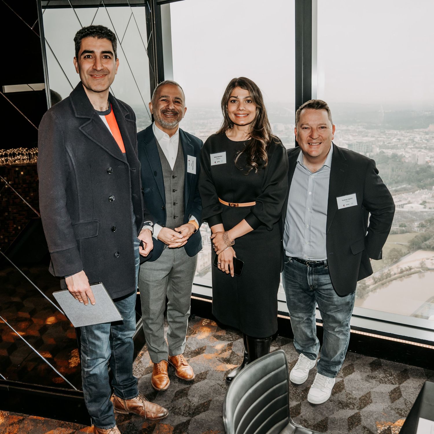 A group of business professionals pose together at a high-rise venue with city views visible through large windows.