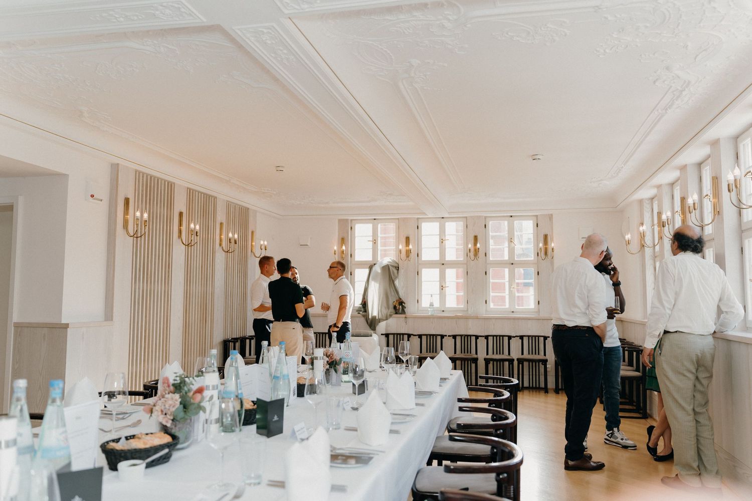 Elegant dining room set for a formal event with long white tables, gold wall sconces, and guests mingling in background.