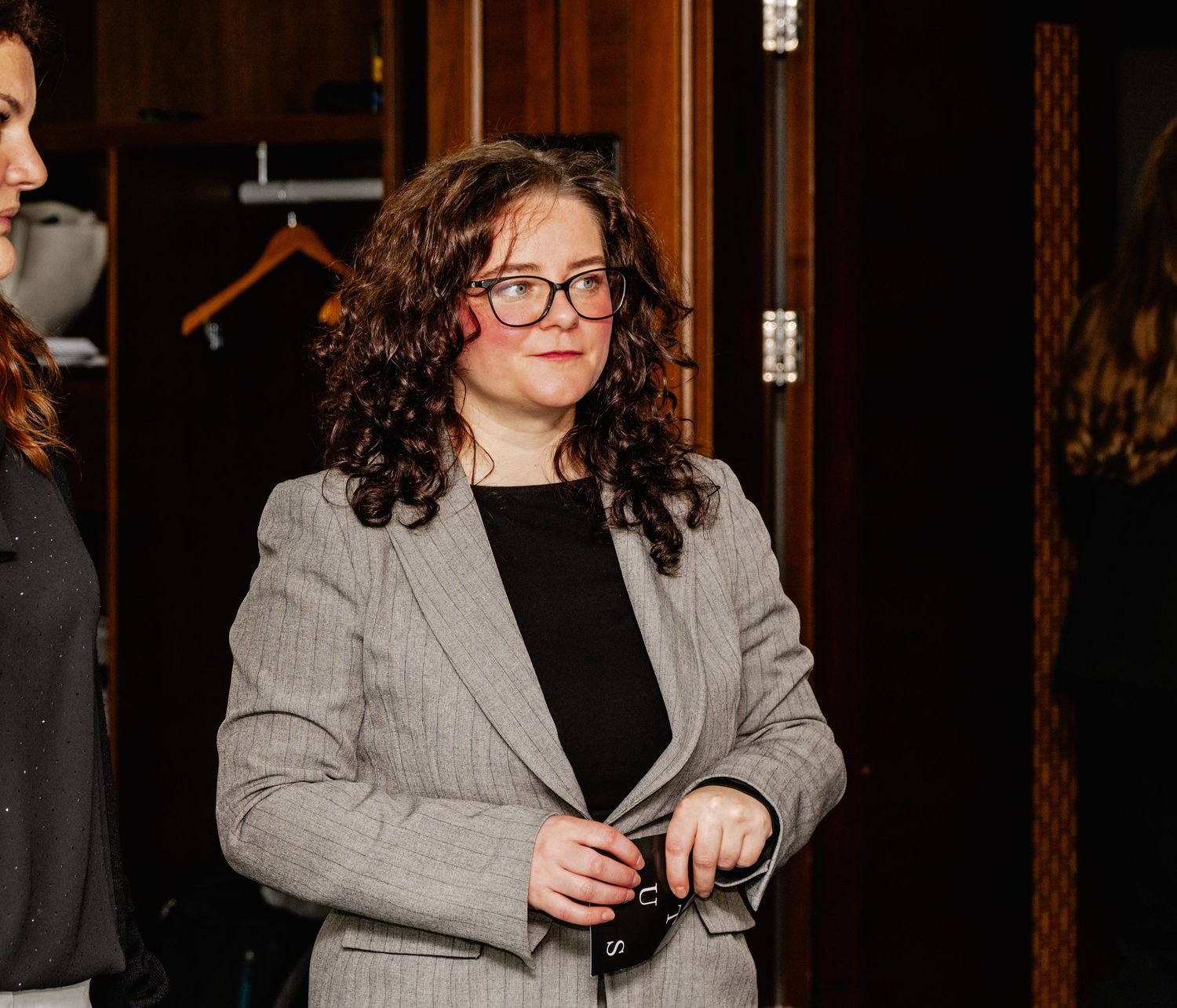 Woman in gray blazer and red glasses holding accessories, standing indoors at a professional event.