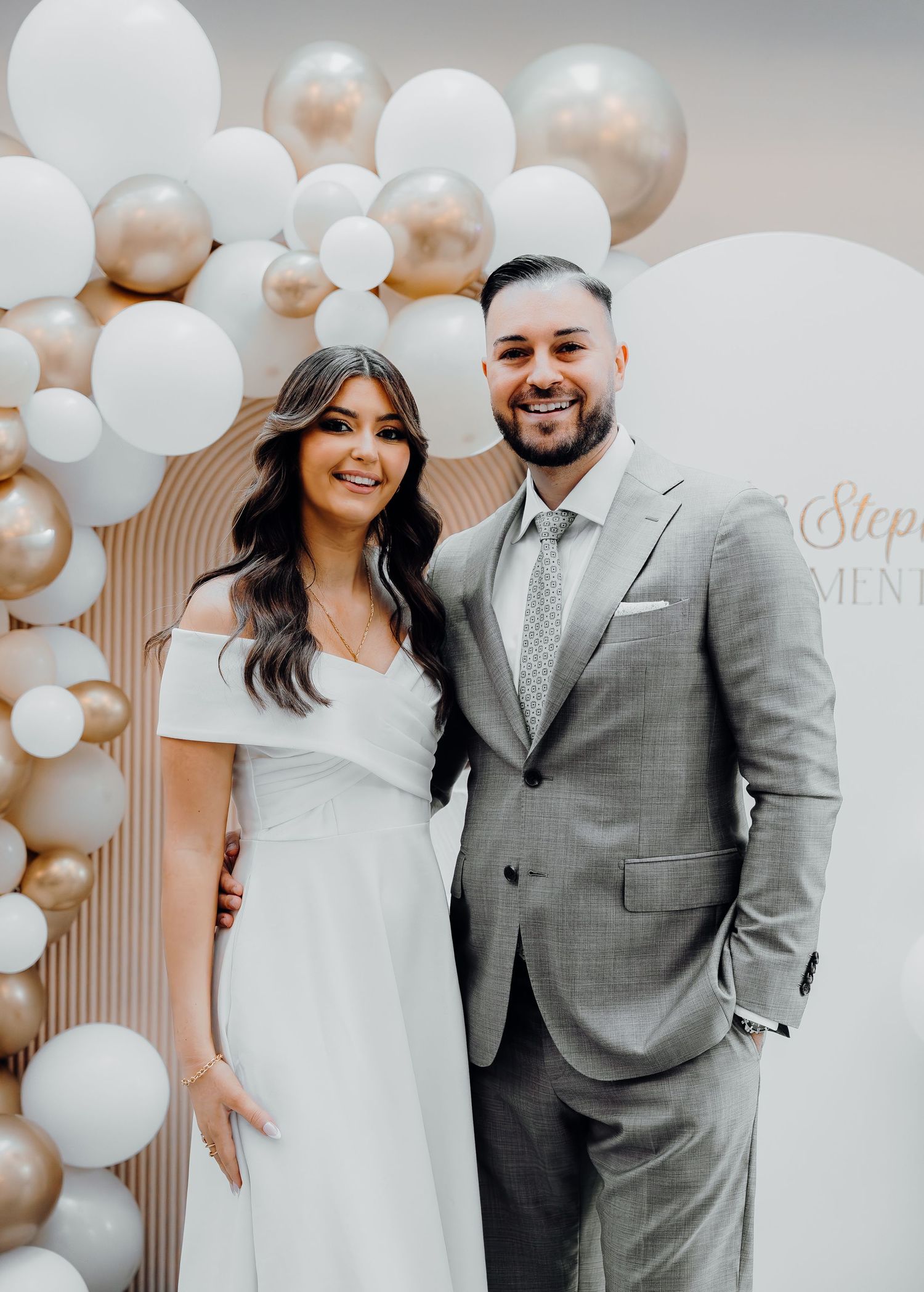 A couple poses together at an elegant event with white balloon decorations and a decorative wall backdrop.