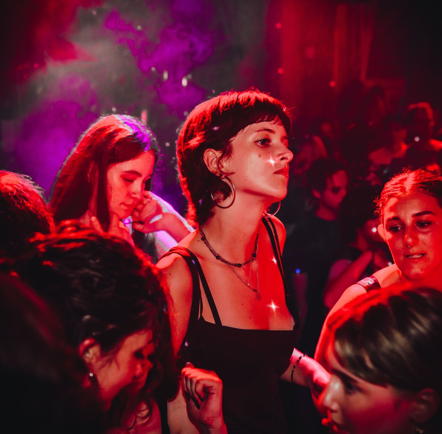 Crowd dancing inside a dimly lit nightclub bathed in dramatic red and purple lighting with dark silhouettes visible.
