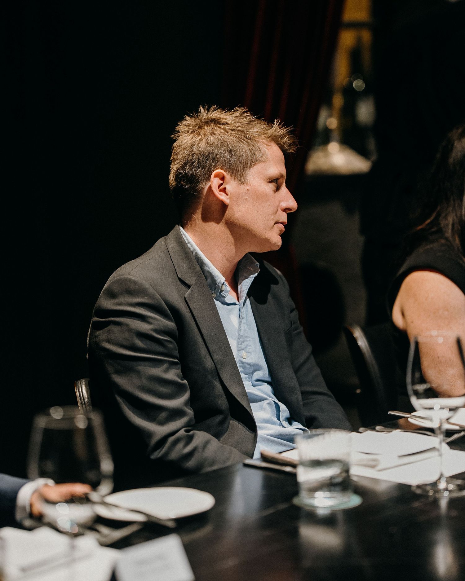 Someone in a dark blazer and light blue shirt sits at a dining table in a dimly lit restaurant setting.