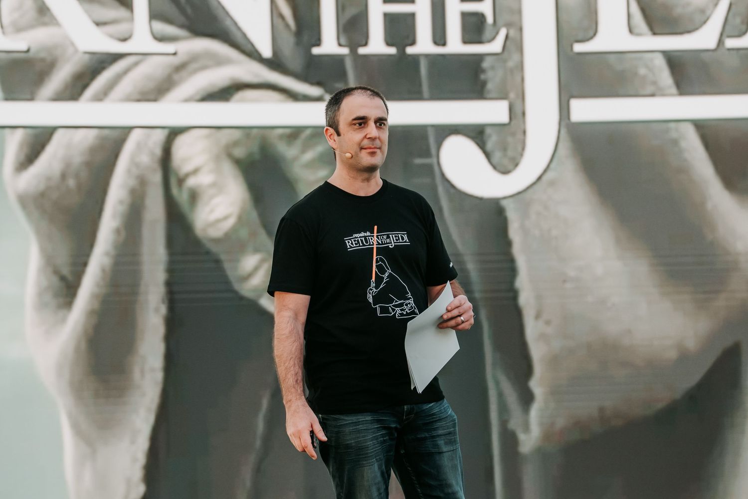 Two people in black t-shirts stand near a wall with text and graphic elements.