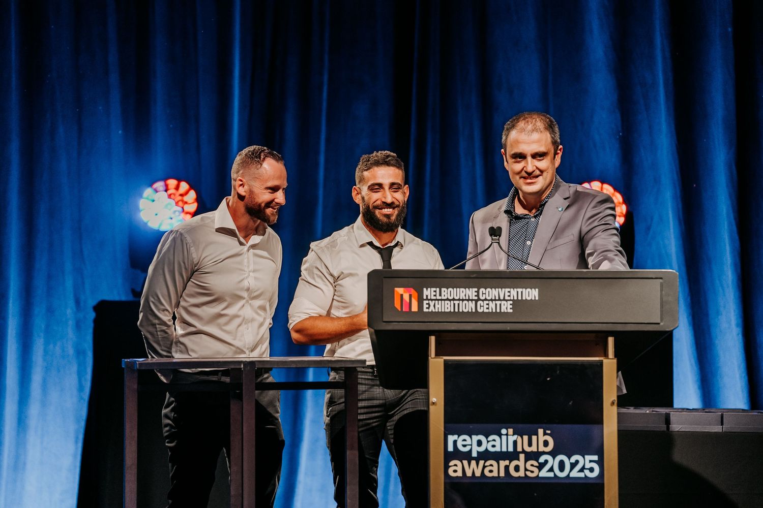 People accepting awards on stage at a business conference with blue curtain backdrop and presentation screen.
