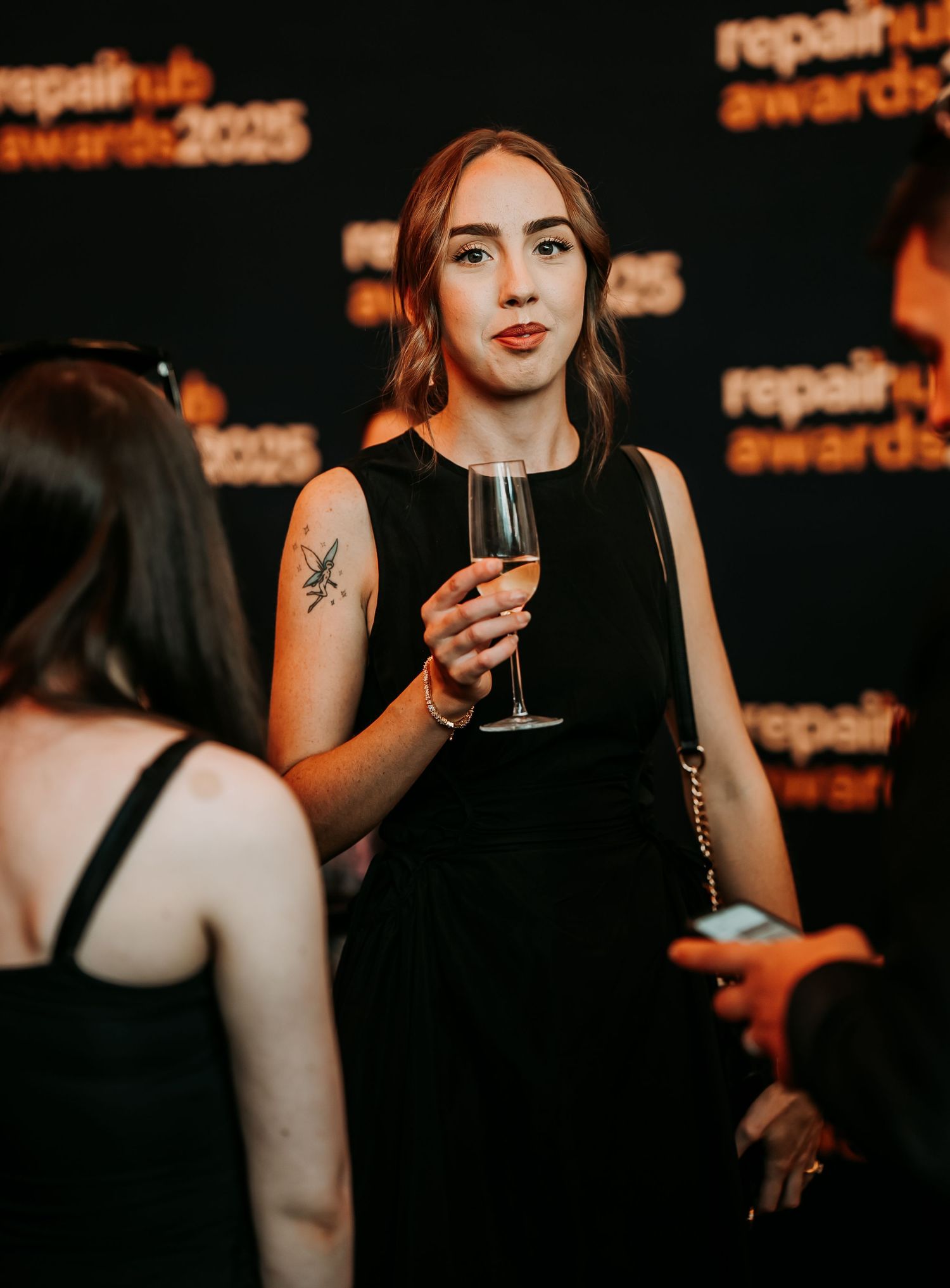 A person in a black sleeveless dress holds a wine glass at an evening event with orange lighting in the background.