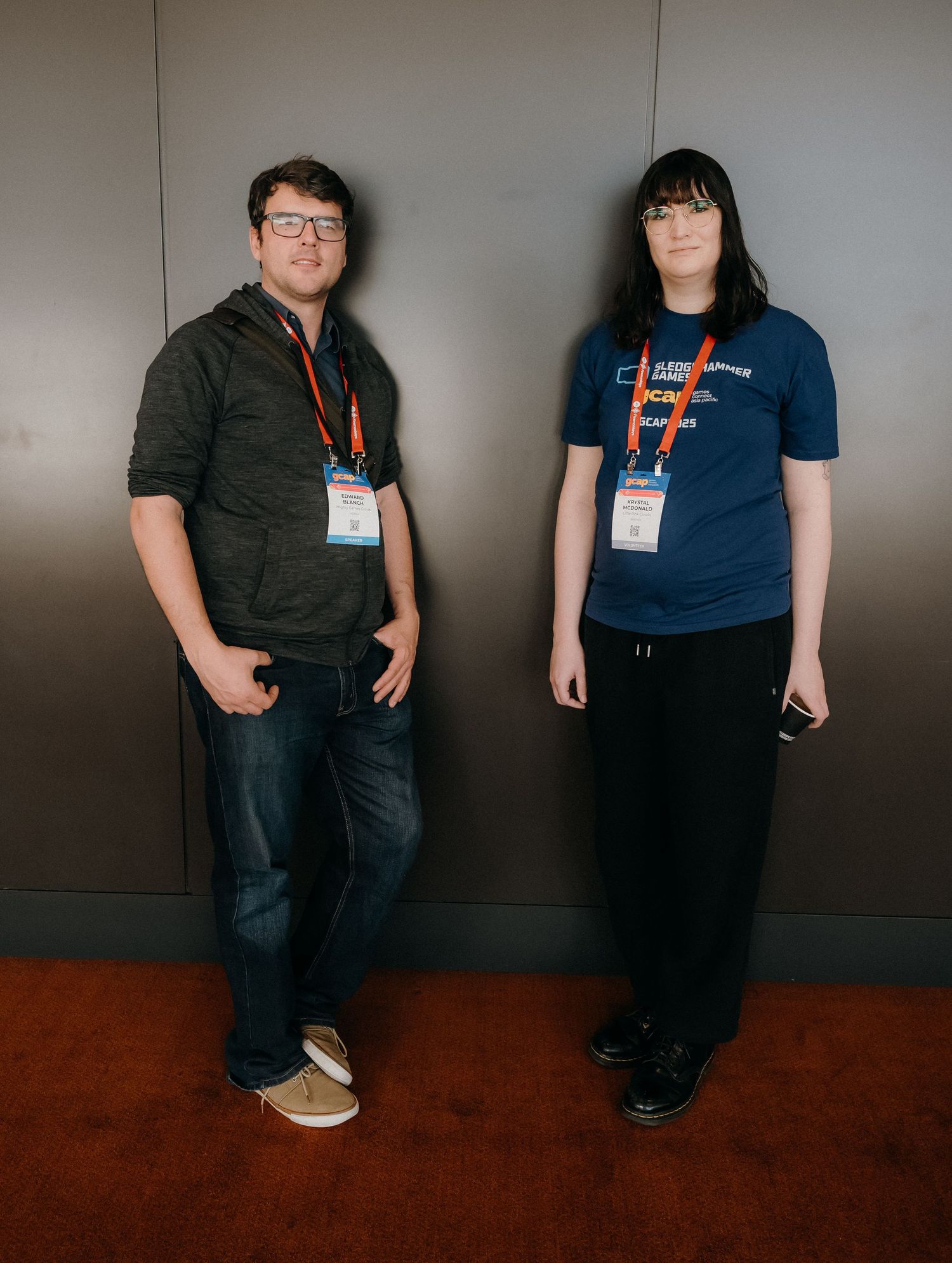 Two conference attendees wearing name badges stand against a gray wall in business casual attire.
