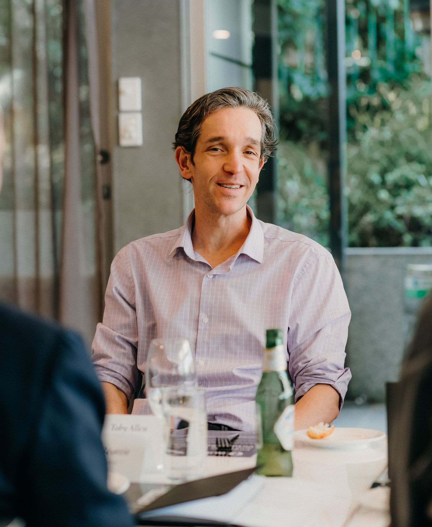 A person in a light colored shirt smiles during a conversation at a restaurant table with drinks.