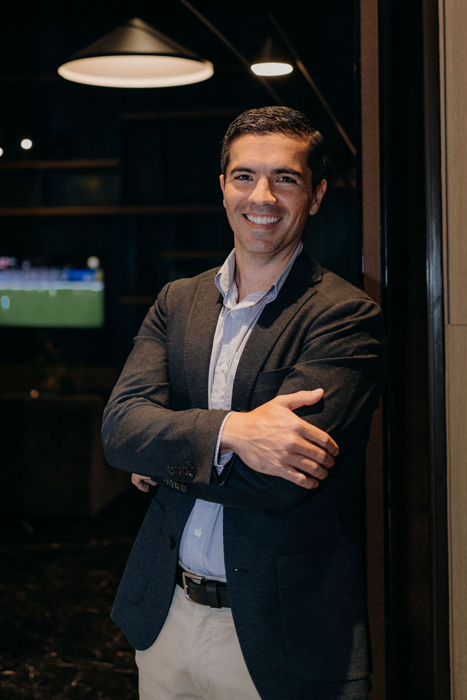 A person in a dark suit jacket stands against a black background with overhead lighting creating dramatic shadows.