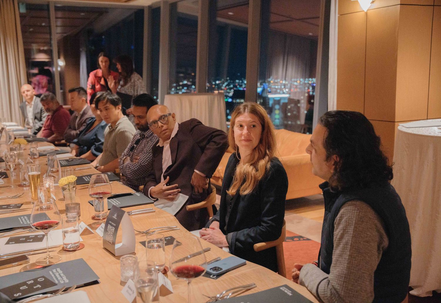 A group gathers around a long table for a business dinner meeting in a warmly lit restaurant setting.