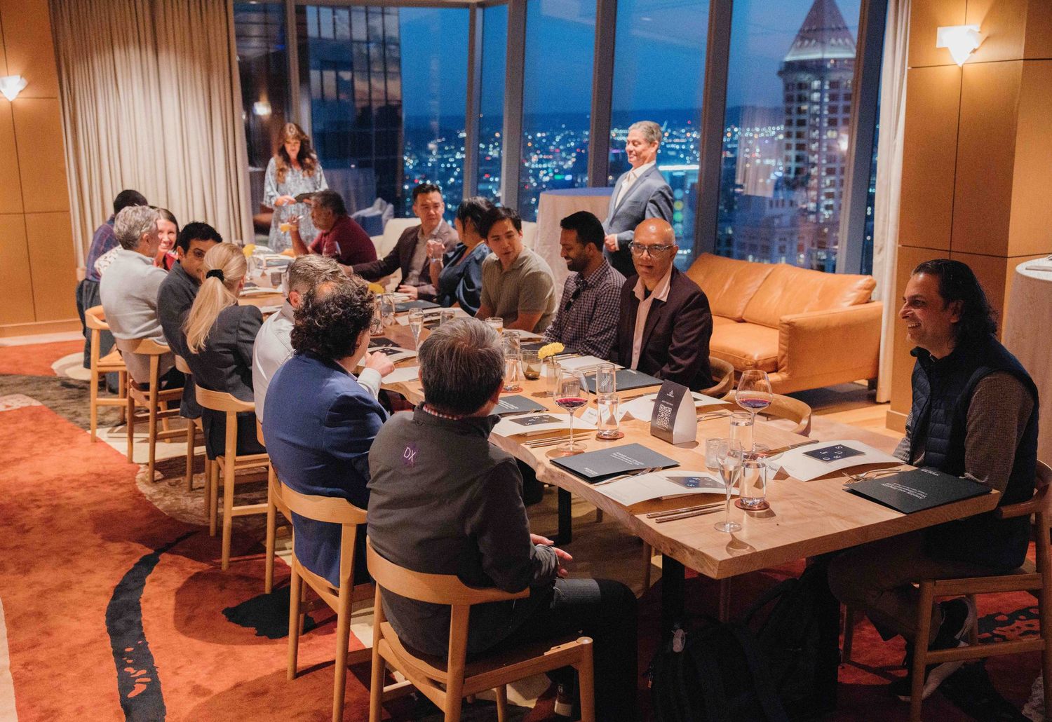 Panoramic view of a modern restaurant dining room with wooden tables and warm lighting during evening service.