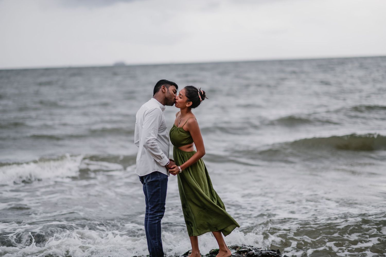 A couple shares romantic moments along a sandy beach with crashing waves in the background.