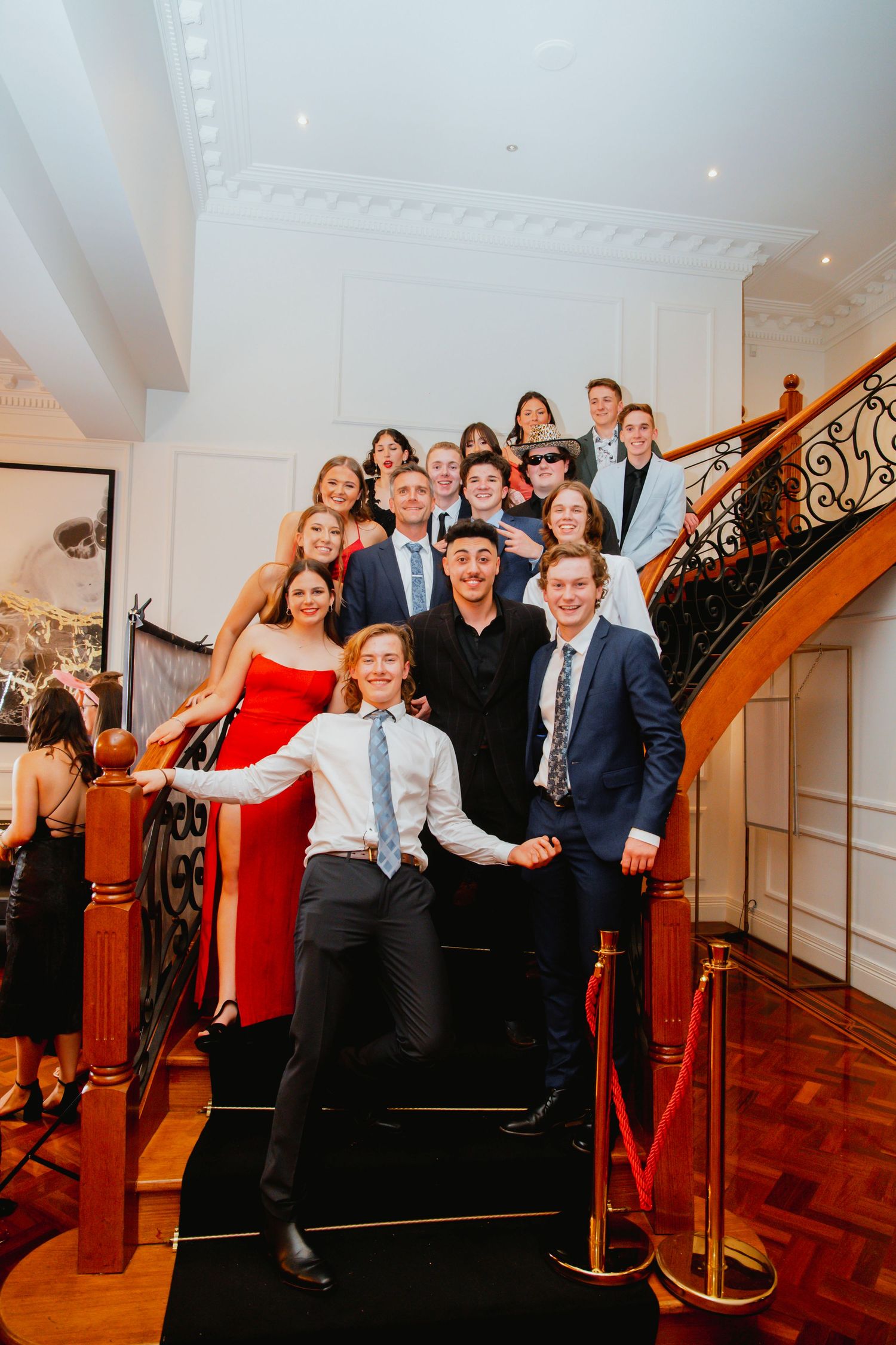 A formal group photo of wedding guests gathering on an elegant wooden staircase with ornate railings.