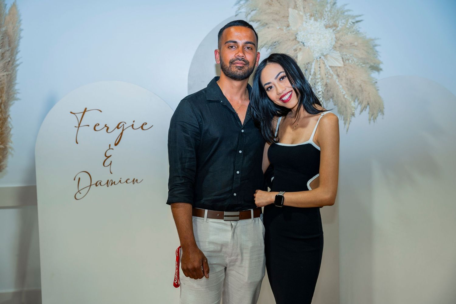 A couple poses at a professional photoshoot against a beige wall with champagne colored palm fronds decor.