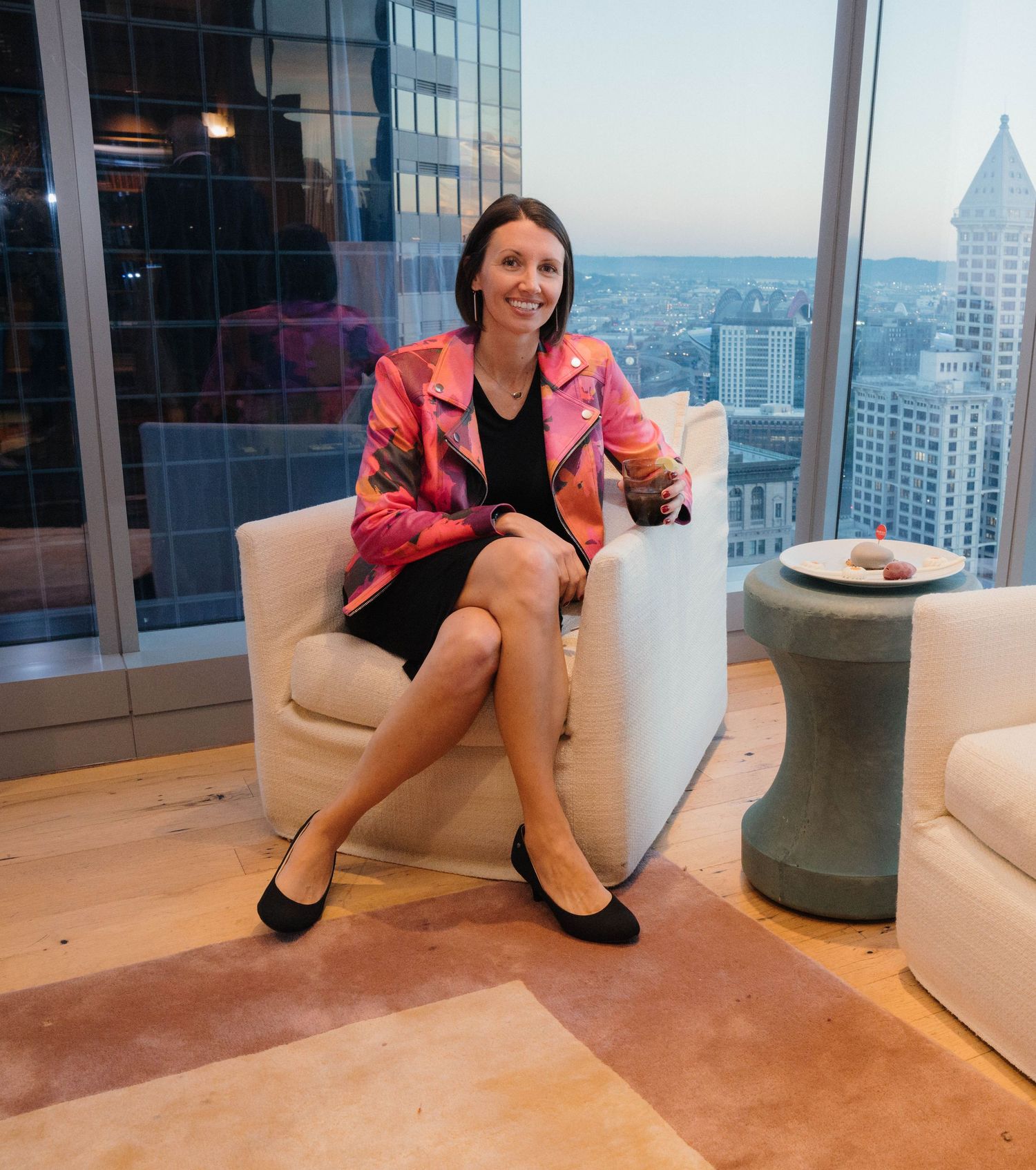 A person in a pink blazer sits on a white couch in a modern high-rise office with city views through large windows.