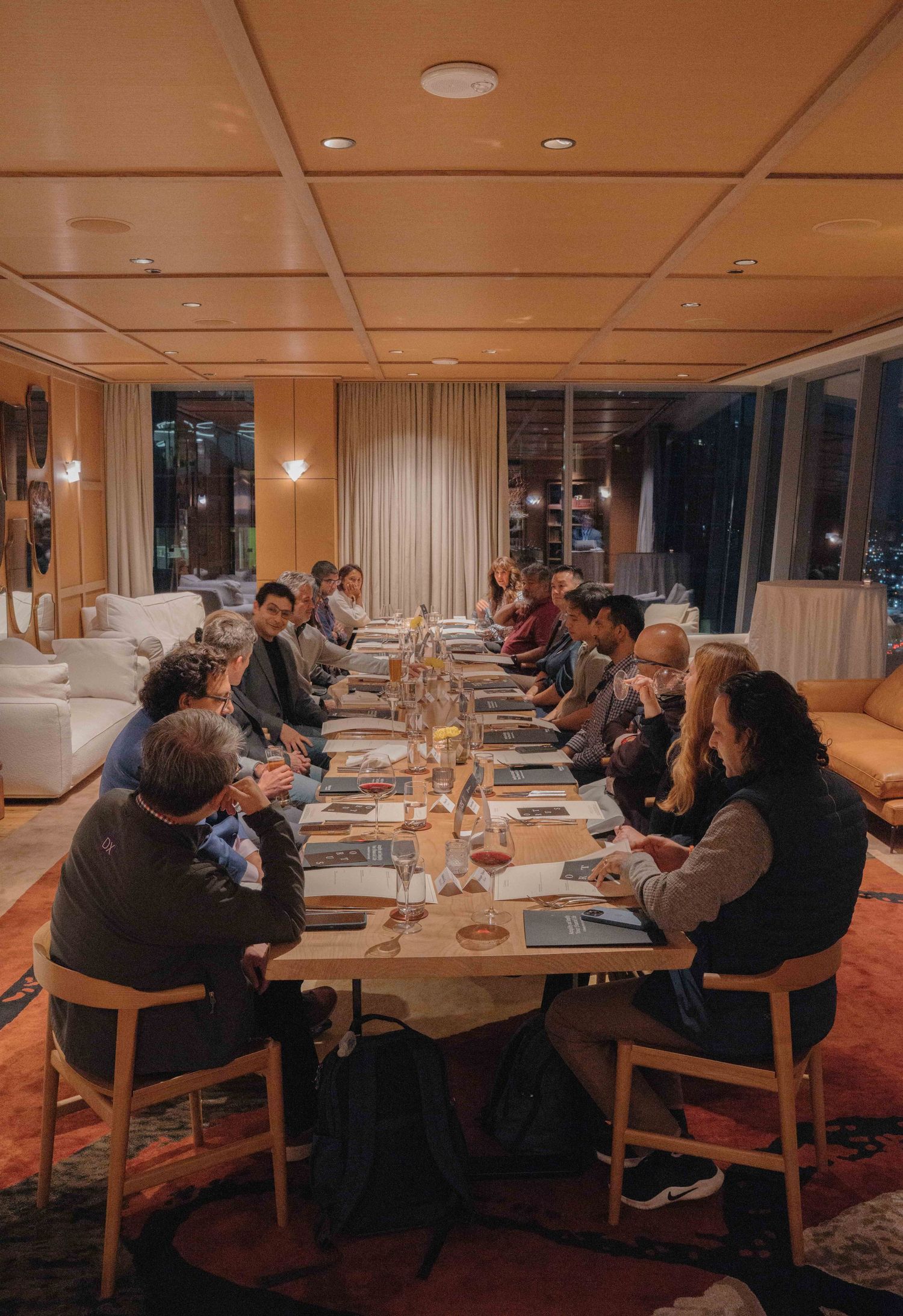 A group gathers around a long dining table in an elegant restaurant with warm lighting and modern decor.