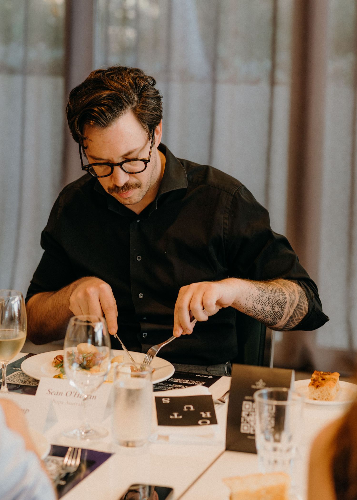 A person wearing glasses and dark clothing dines at an elegant restaurant table with multiple courses and wine glasses.