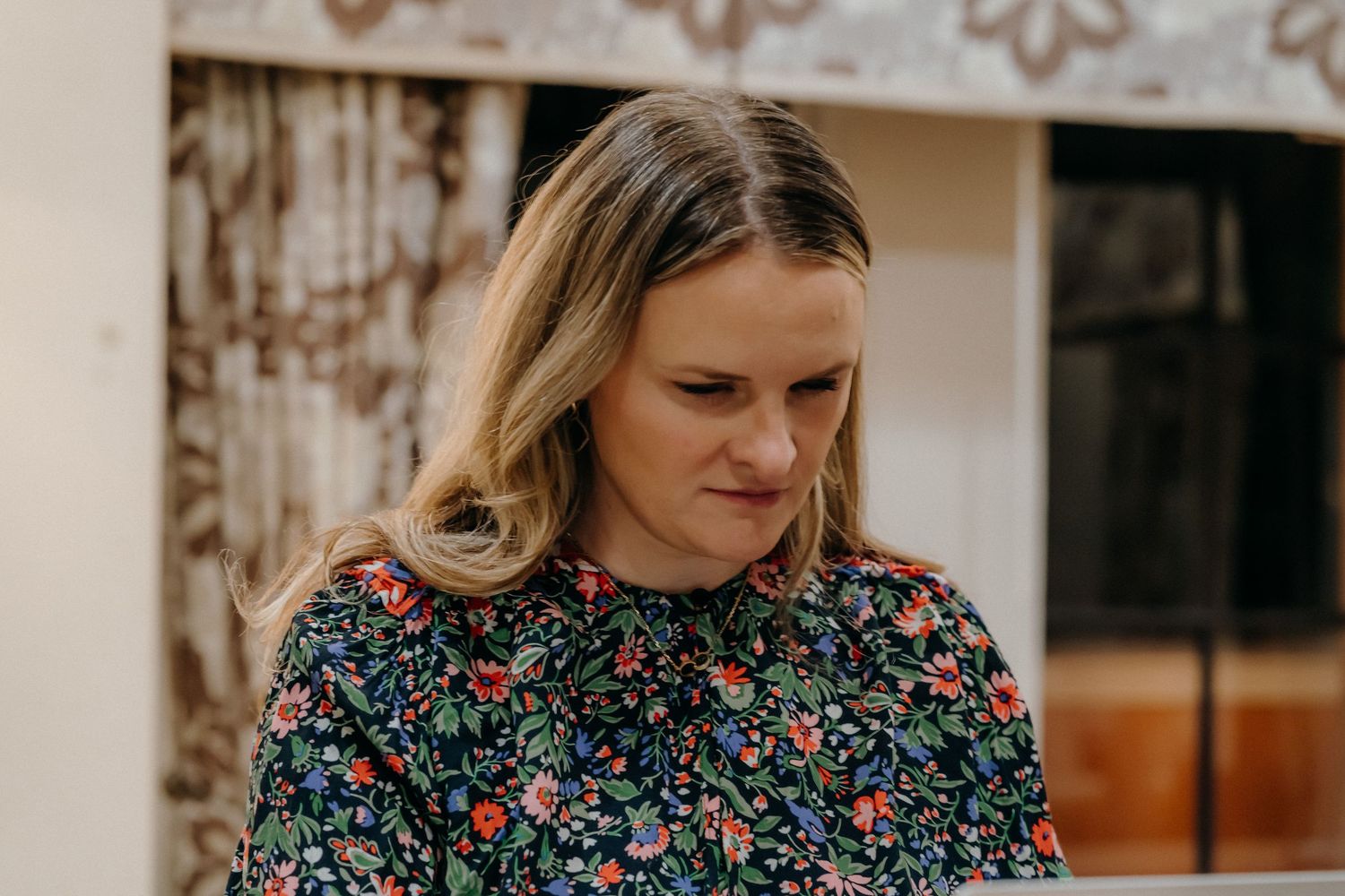 Person in a floral dress stands in a home interior setting with patterned wallpaper in the background.