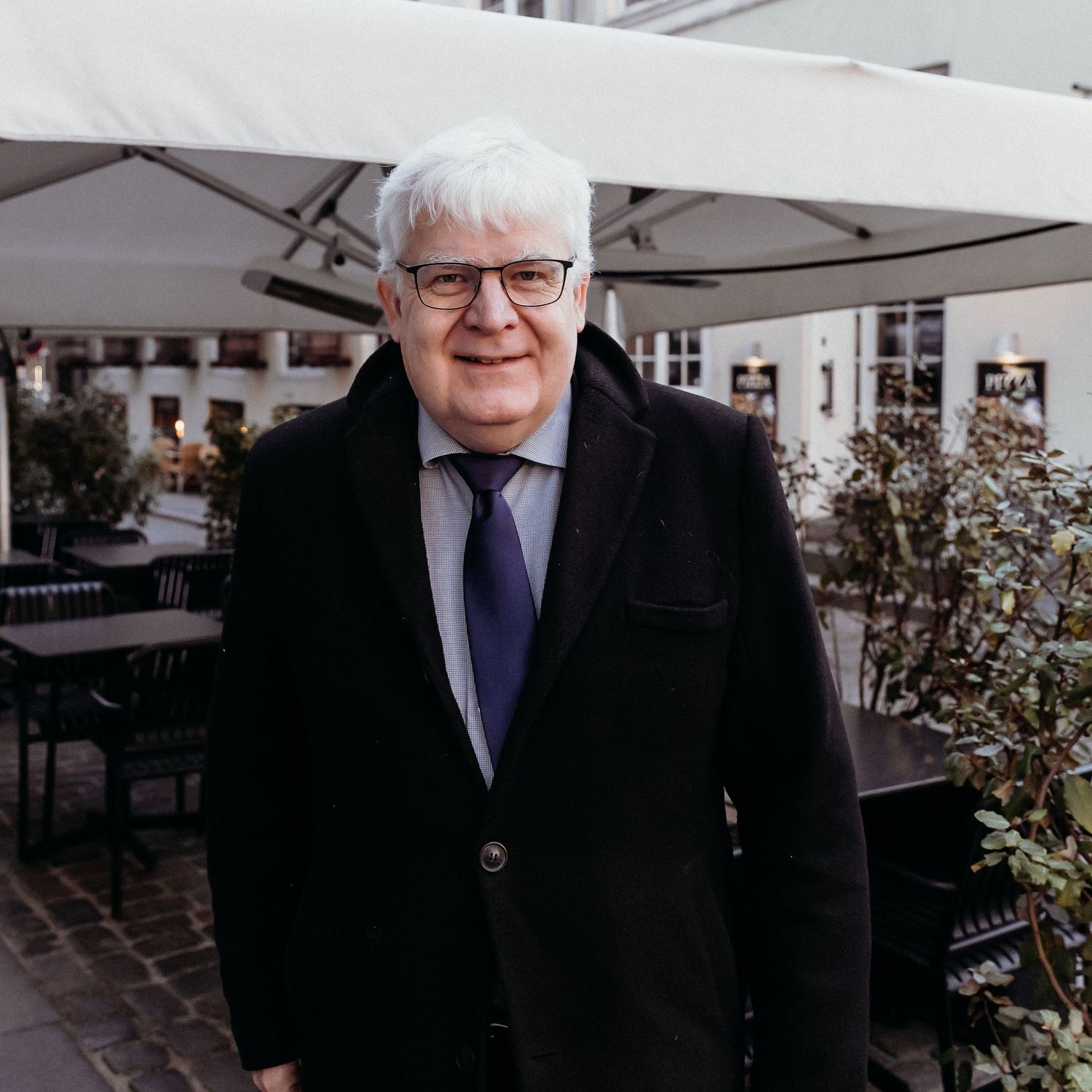 Person in a dark suit and glasses stands outdoors near white patio umbrellas and foliage.