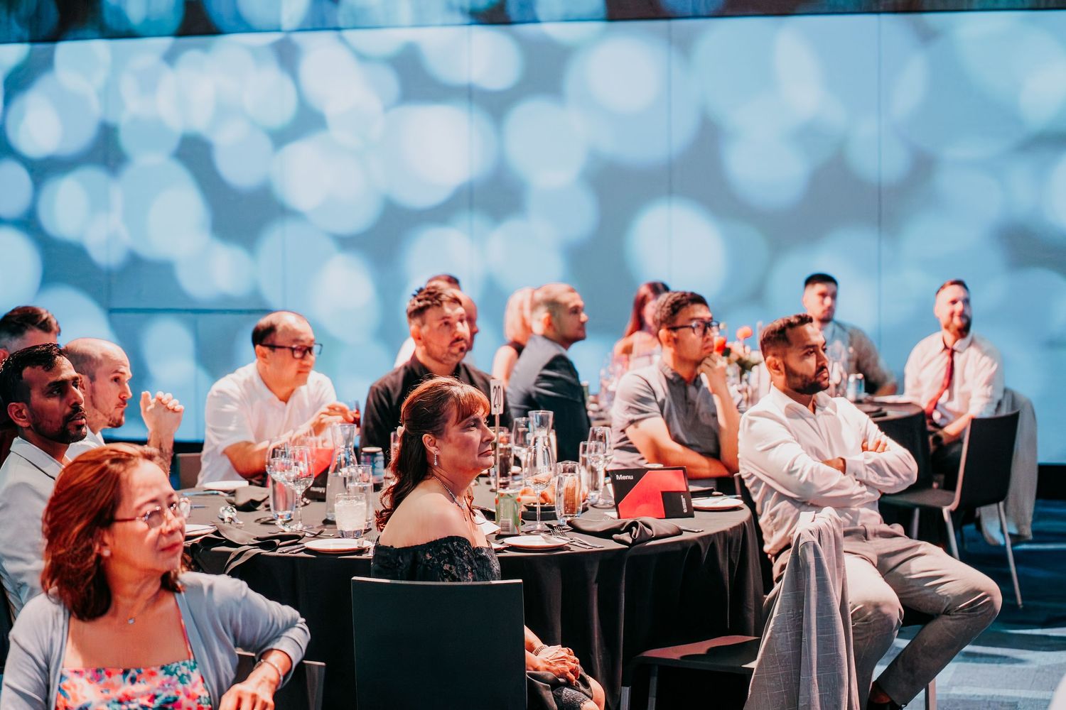 Event attendees seated at round tables with blue bokeh lighting effects in the background during a social gathering.