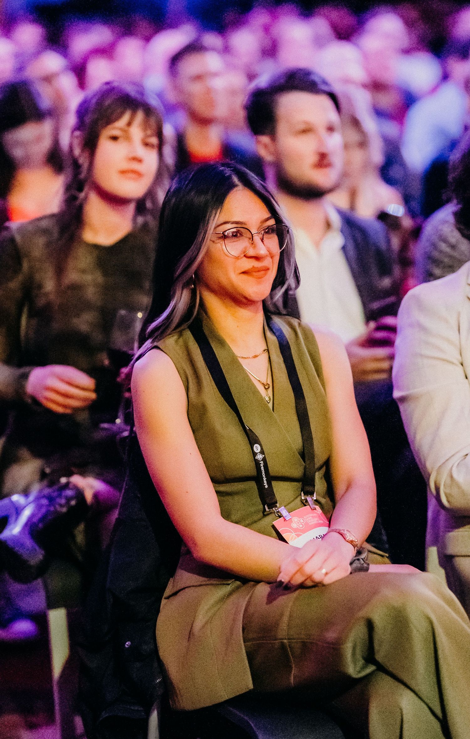 Person in olive green sleeveless dress sits attentively in audience at crowded indoor event with purple lighting.
