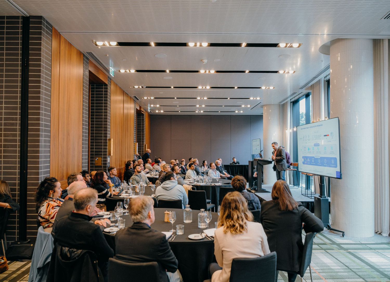Business professionals attend a corporate conference presentation in a modern meeting room with floor-to-ceiling windows.