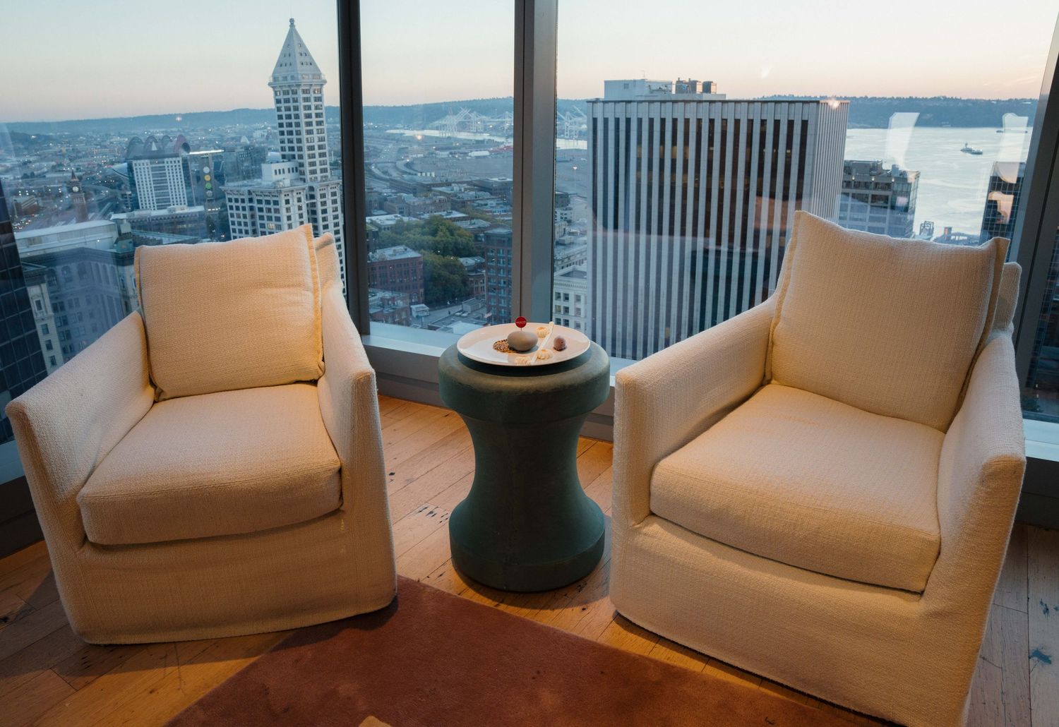 A row of white armchairs arranged in a modern high-rise apartment with city views through floor-to-ceiling windows.